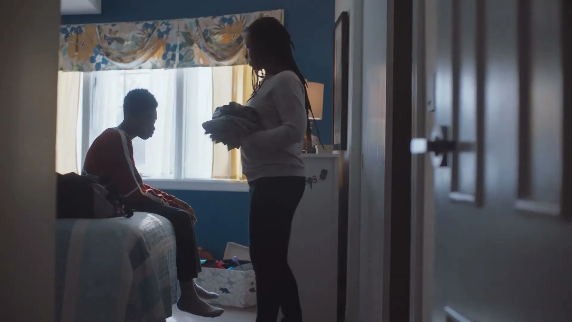 A young man sits on a bed while a woman stands nearby, holding clothes, in a bedroom with a window.