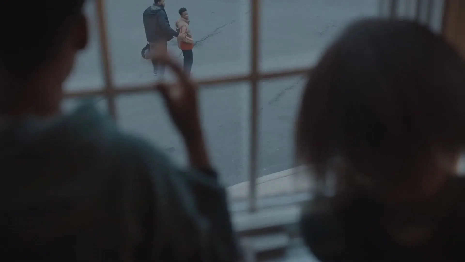 Two children watch from a window as a father and son walk away on a street, the son waving goodbye.