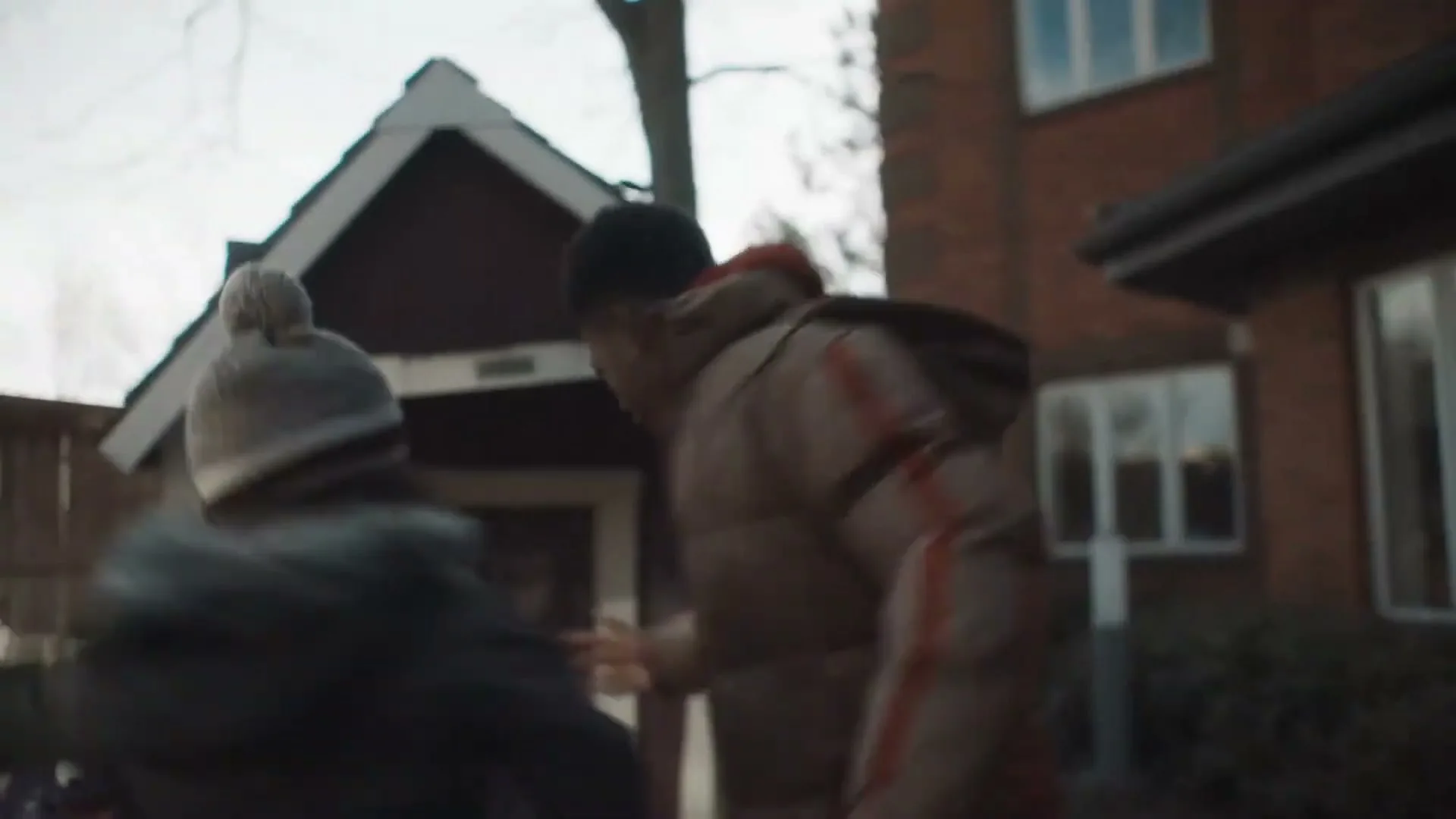 A young male in a puffer jacket and hoodie runs past a small red shed and a brick building, with another person briefly visible.