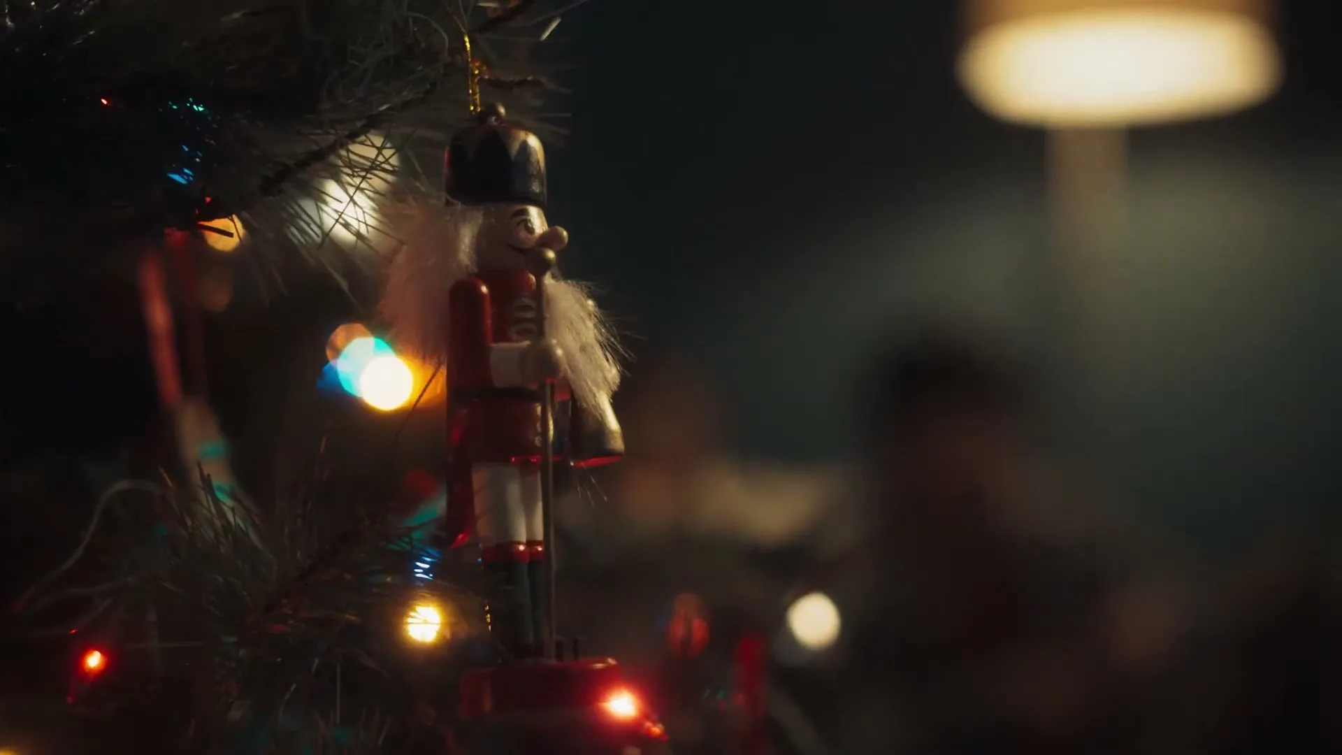 A close-up shot of a nutcracker ornament hanging on a Christmas tree, with blurred lights and a lamp in the background.