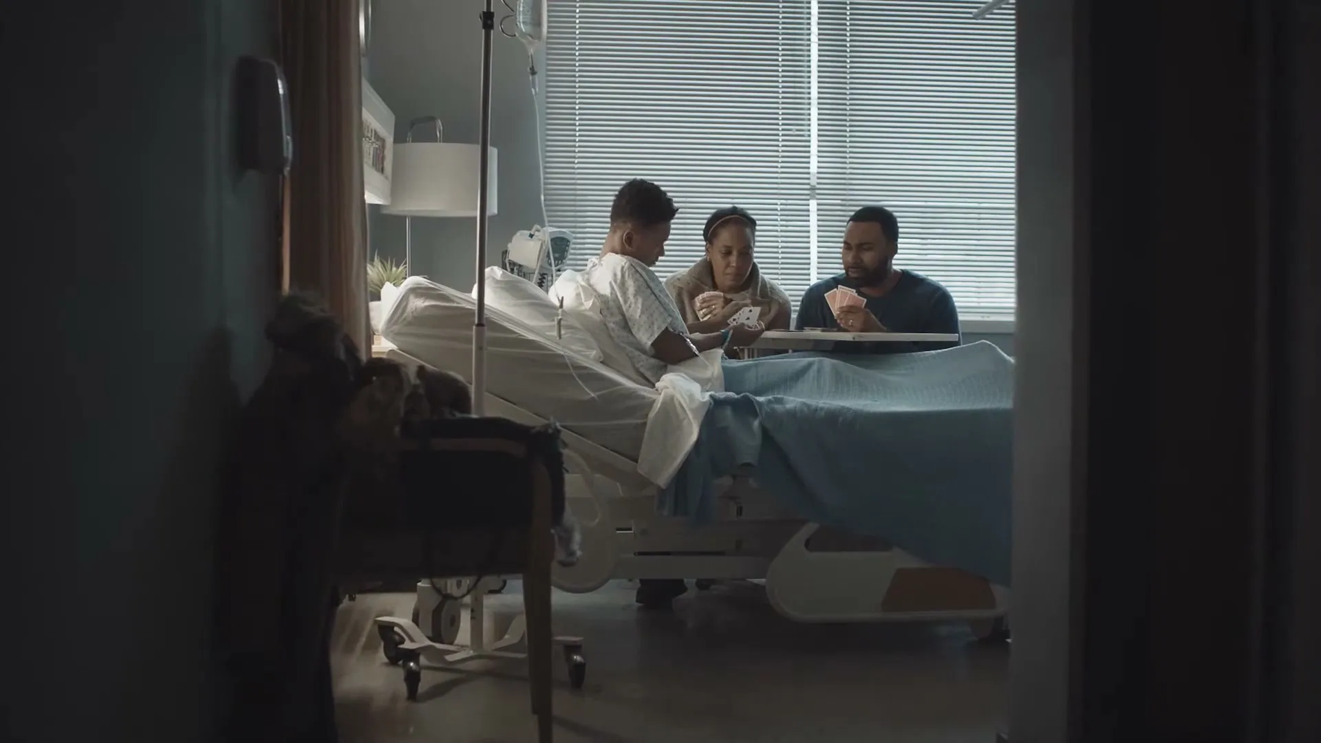 A young boy in a hospital gown plays cards with a woman and a man at his bedside in a hospital room.