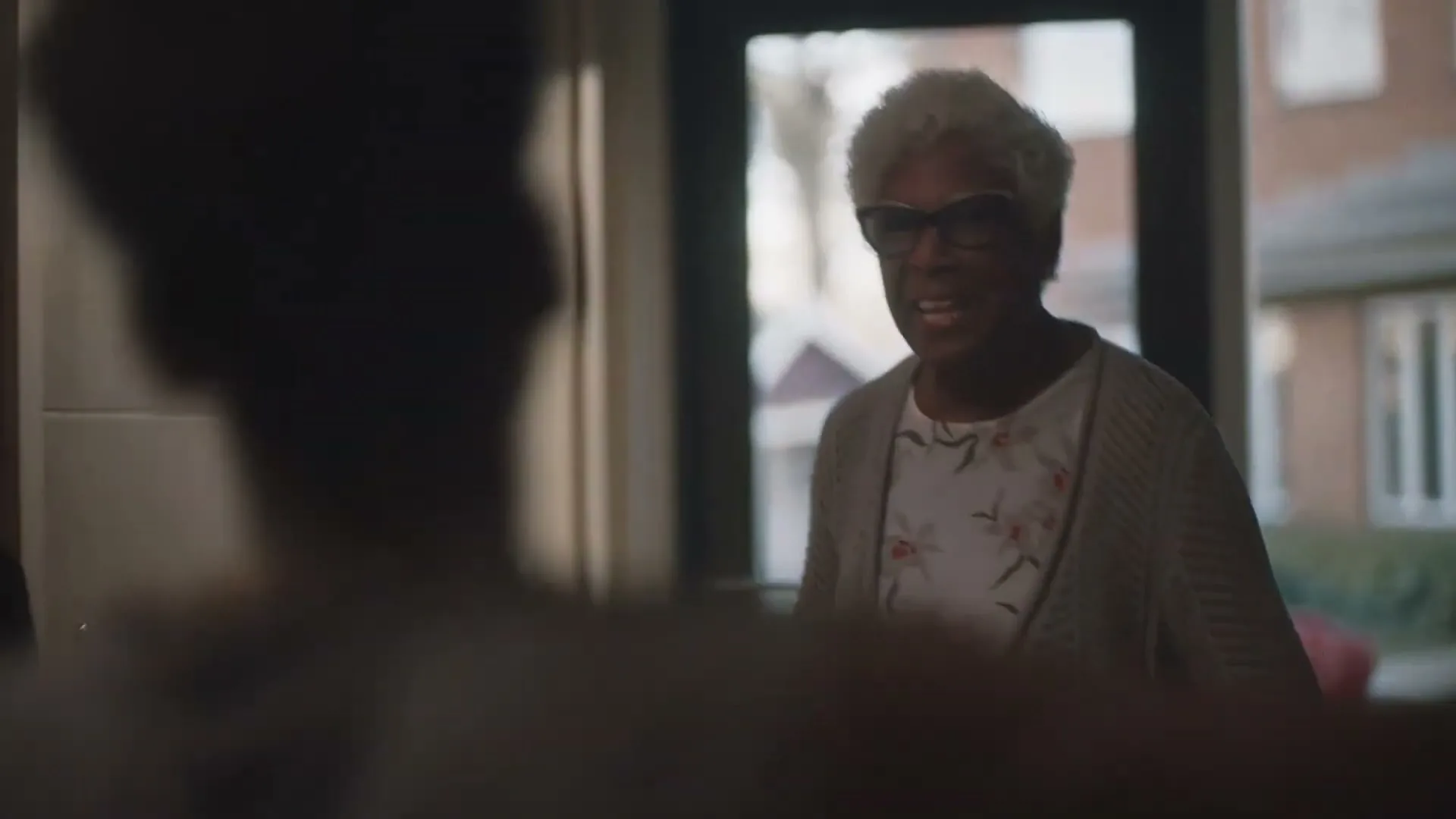 An older Black woman with short white hair and glasses stands in a doorway, smiling warmly at someone out of frame, as she greets them.