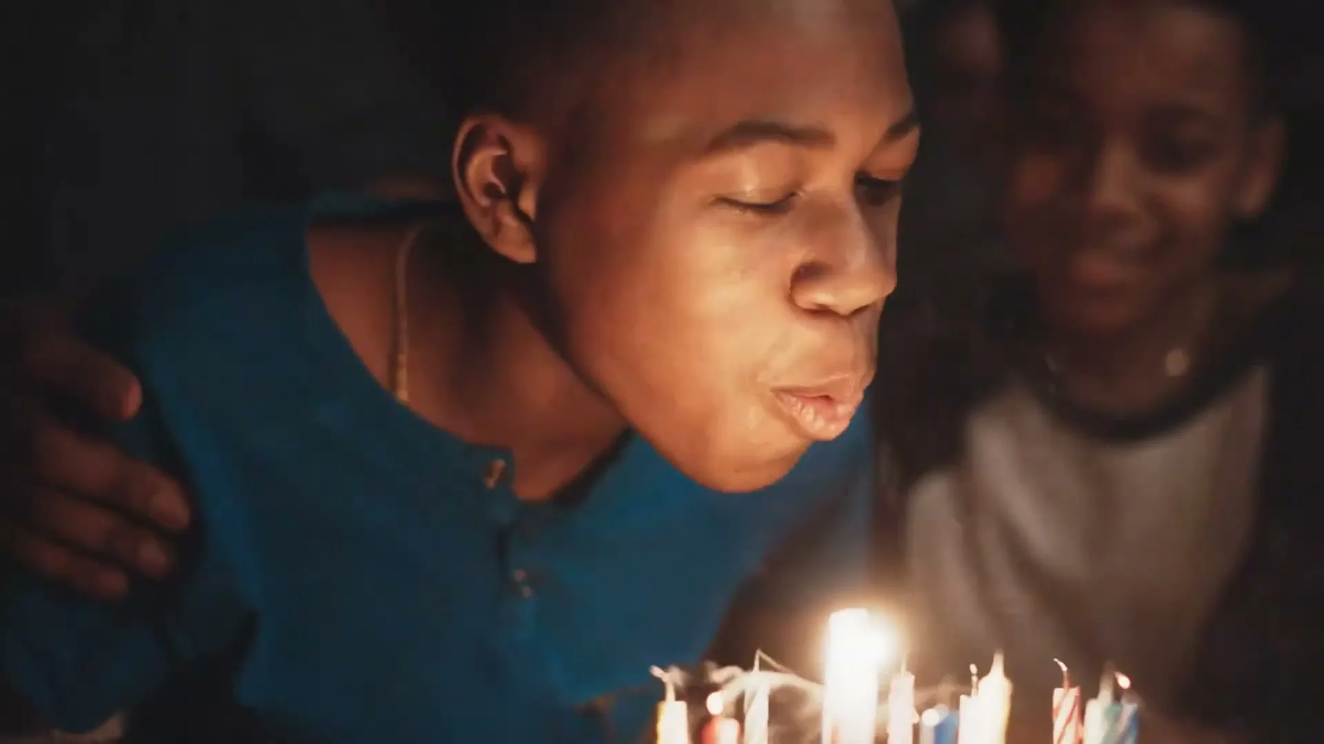 A young Black boy in a blue shirt blows out birthday candles on a cake, with another boy smiling behind him in a dark, intimate setting.