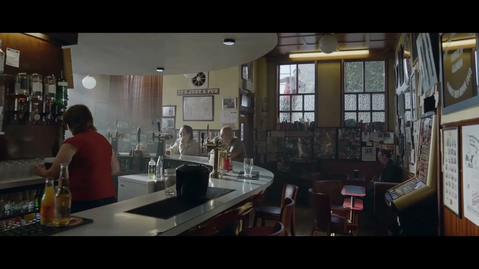 A female bartender in a red top wipes down the bar in a dimly lit pub, while a few patrons sit at tables and the bar.
