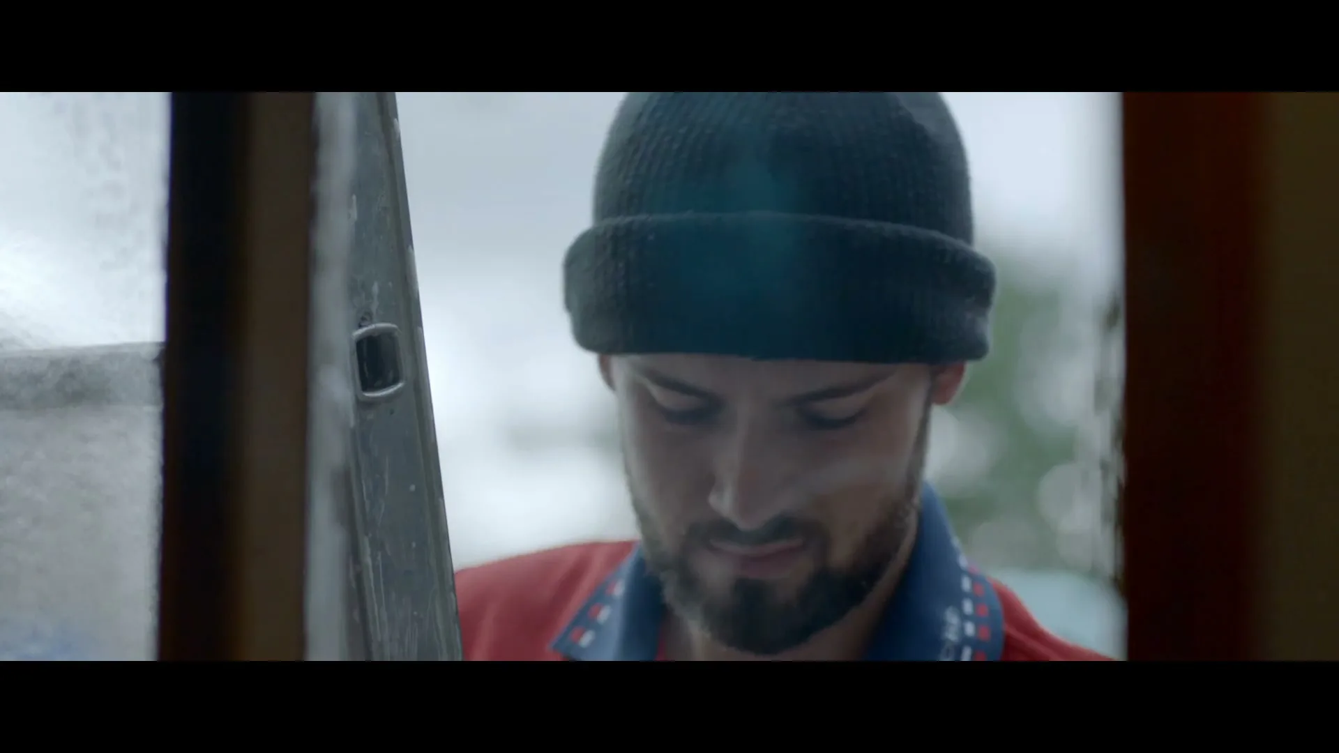 A man with a beard and a black beanie looks out a window, then looks up, in a close-up shot.