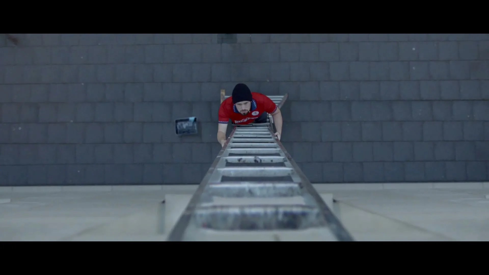 A young man in a red shirt and black beanie climbs a ladder, viewed from directly above, against a grey tiled wall.
