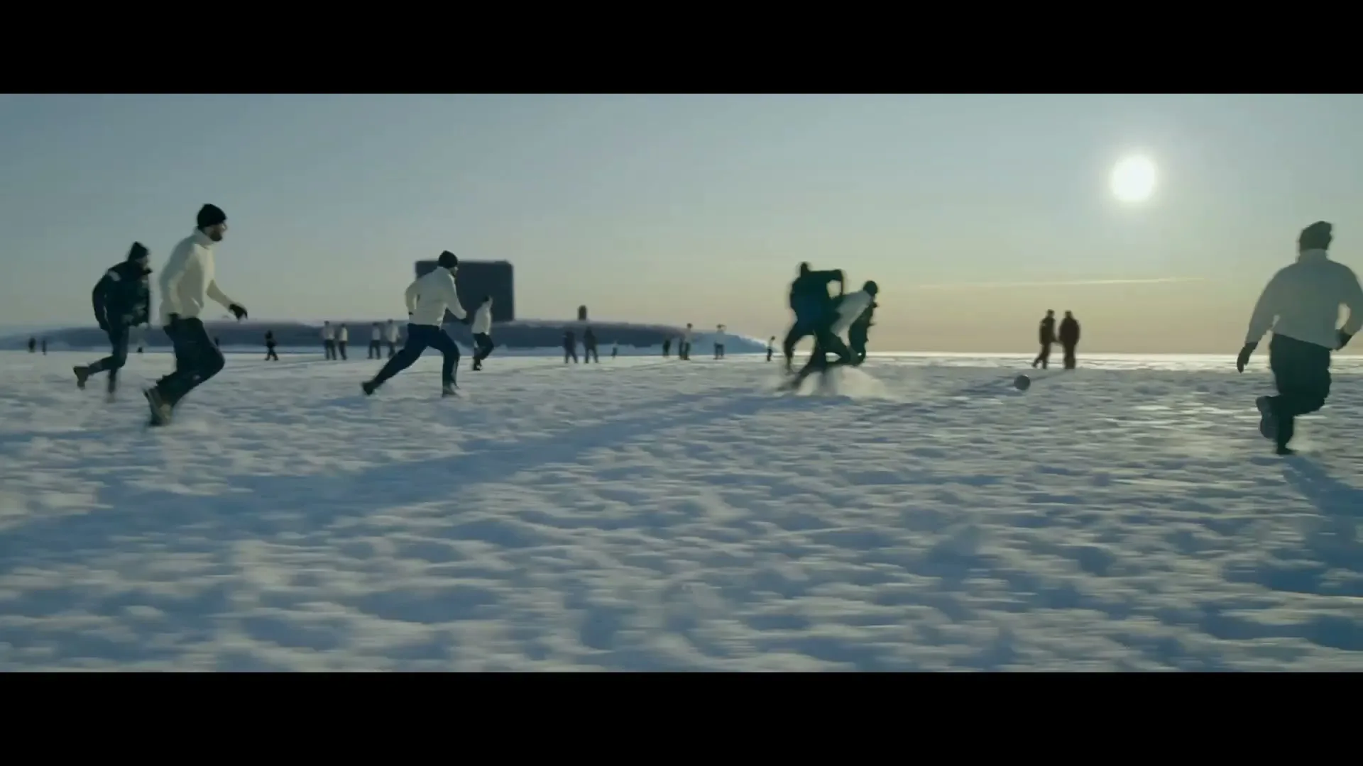 A group of men in winter clothing play soccer on a vast, snow-covered plain with a submarine in the background under a bright sky.