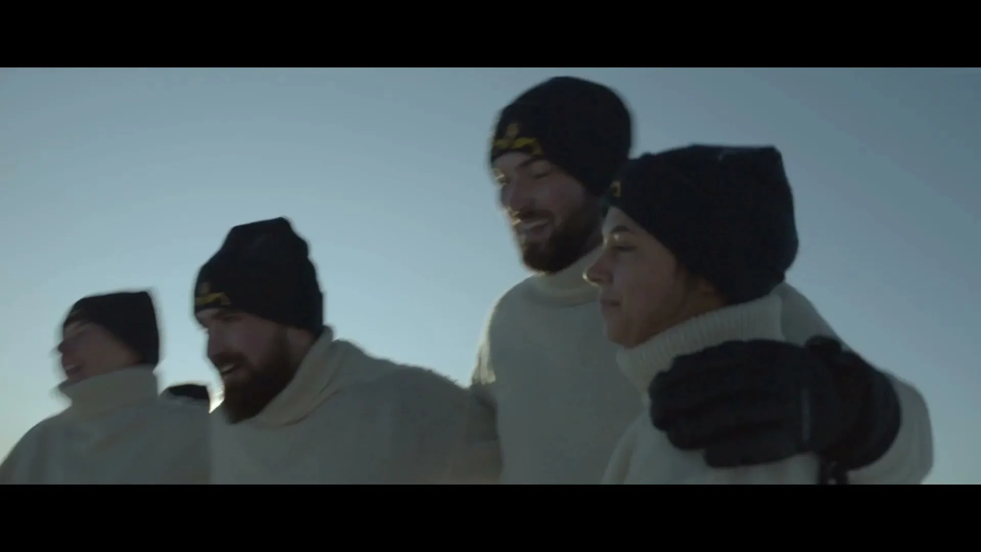 A small group of four young adults, wearing white sweaters and black beanies, are laughing and walking together against a clear blue sky.