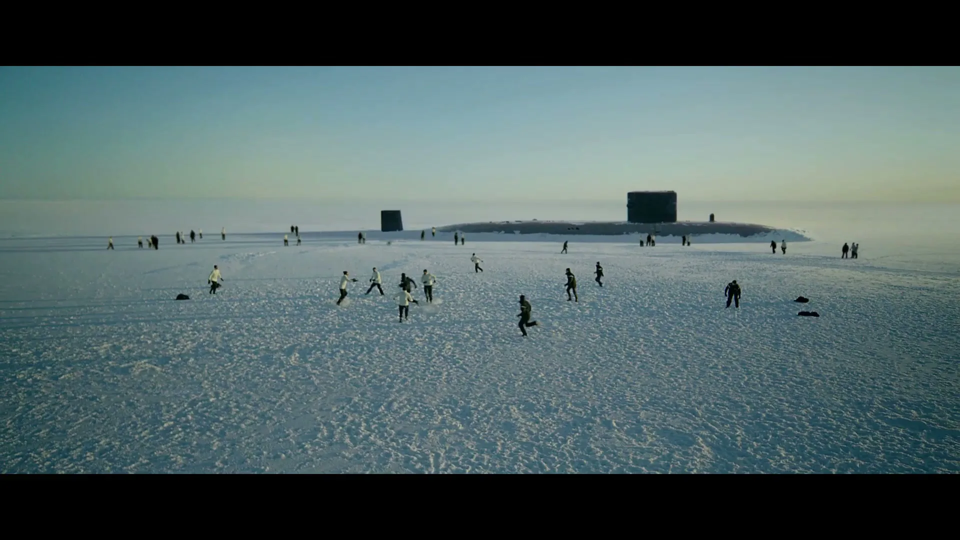 A wide shot shows a group of people playing football on a vast, snow-covered ice field with two submarines visible in the background under a clear sky.