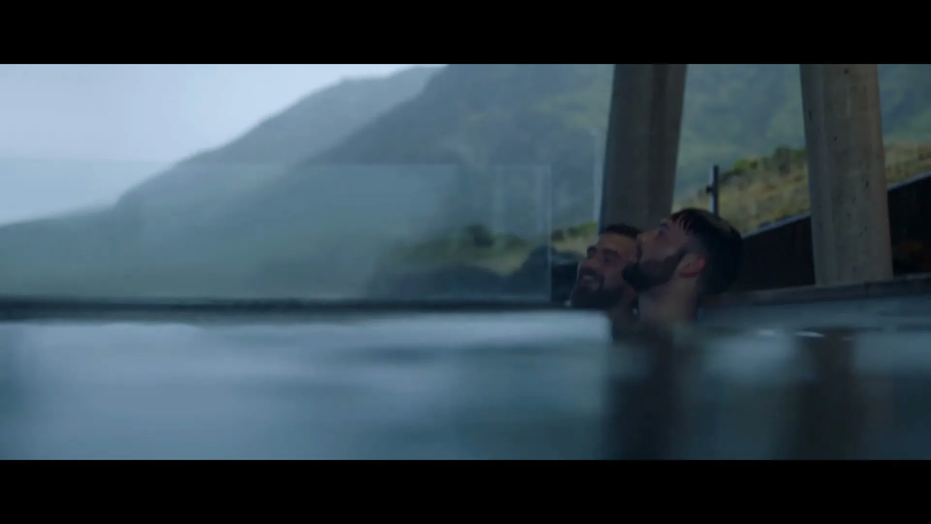 Two men with beards relax in a hot tub outdoors, with a misty mountain landscape in the background.