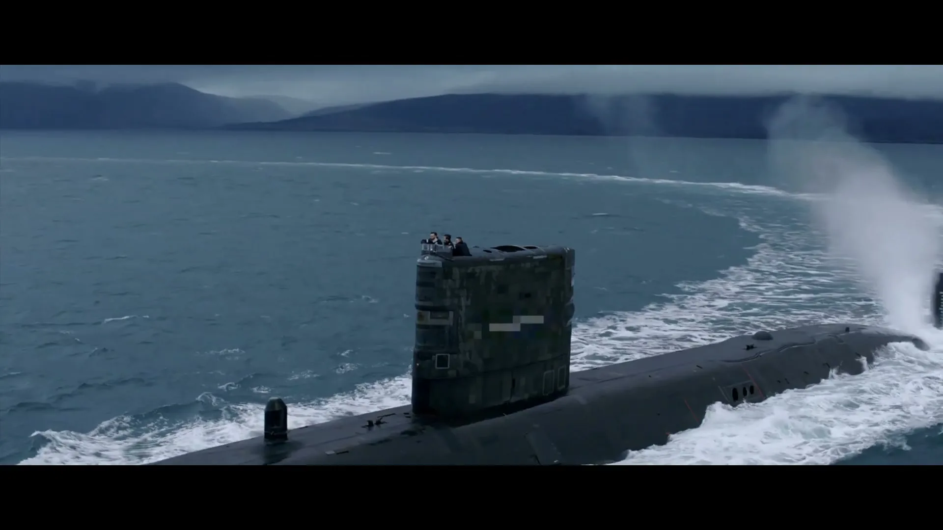 A Royal Navy submarine with a small group of submariners on its conning tower moves through choppy waters with mountains in the background.