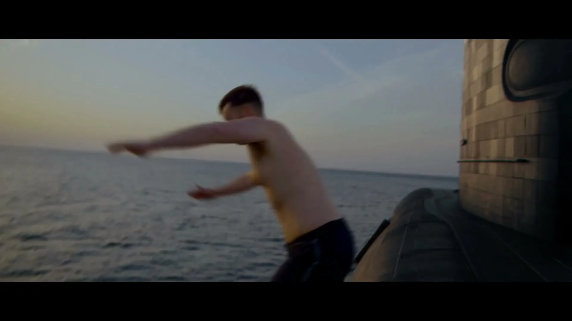A young man in swim shorts stands on a submarine at sea, then jumps into the water, creating a large splash.
