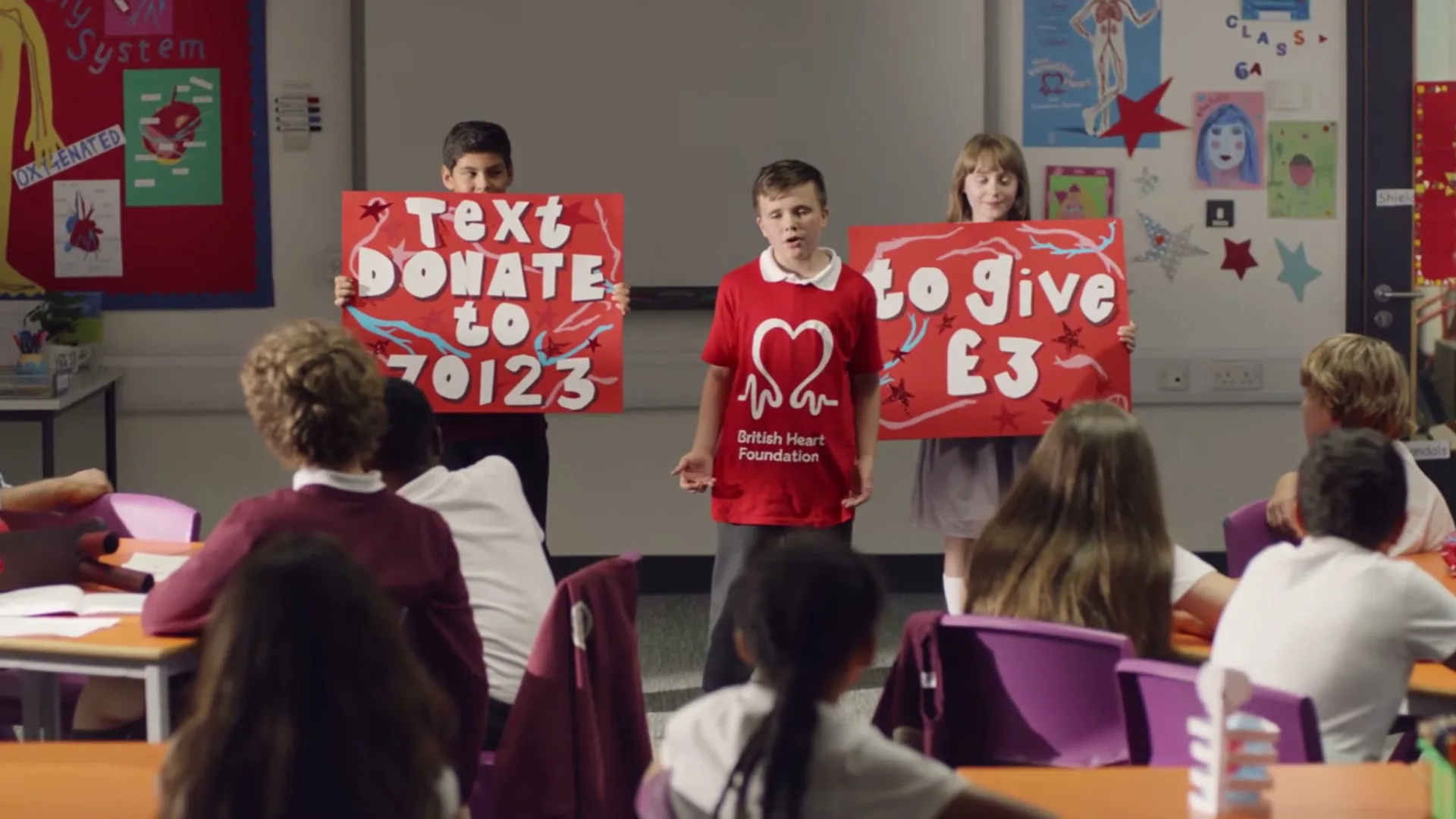 Three children stand at the front of a classroom, holding red signs with white text, while other students sit at desks.