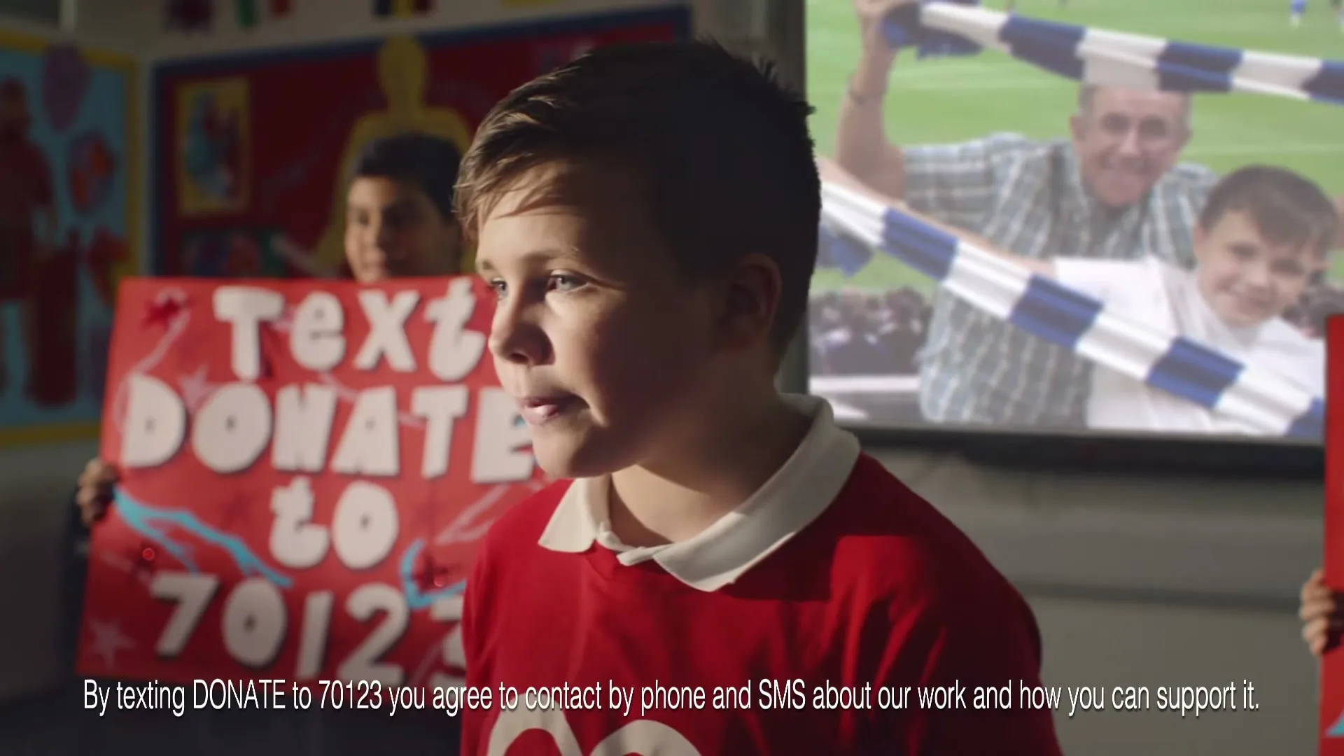 A young boy in a red shirt looks at a screen showing a female doctor, while another boy holds a "Text Donate" sign in a classroom.