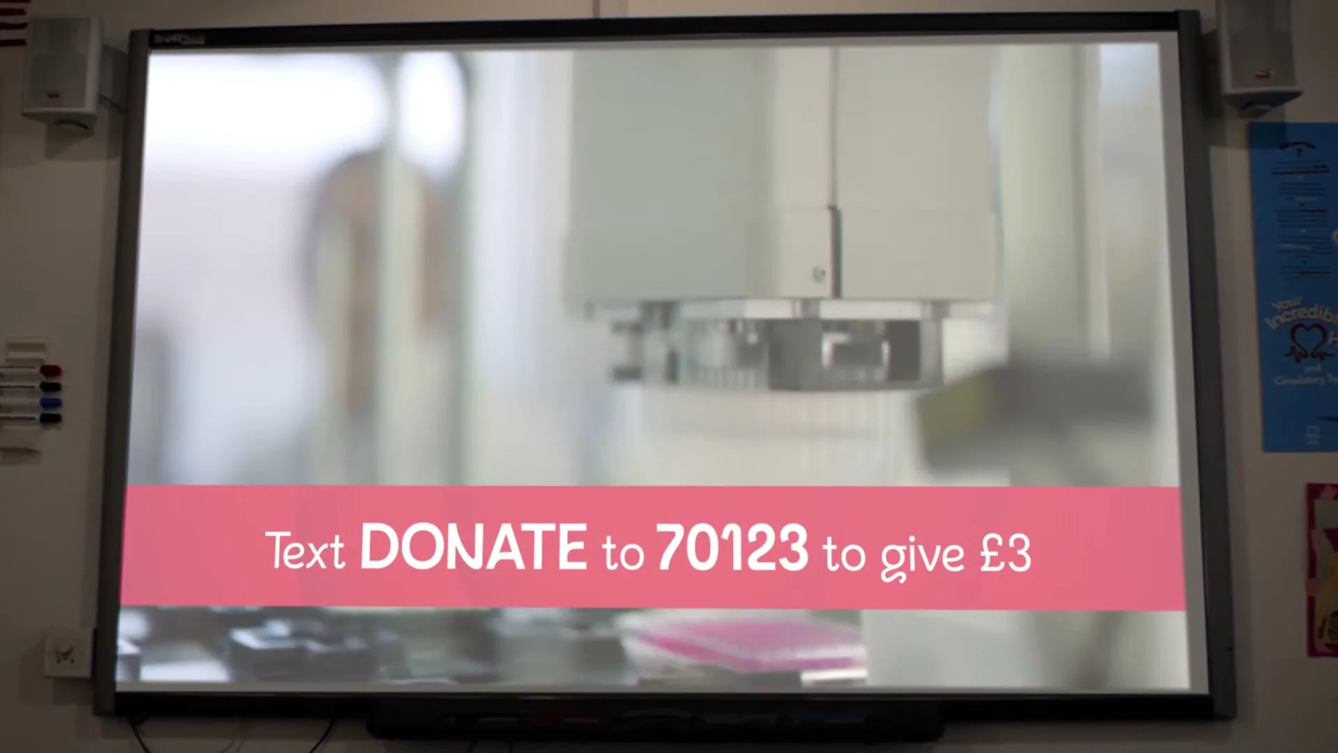 A white interactive whiteboard displays a blurred video of a laboratory machine, with a pink banner at the bottom showing text to donate.
