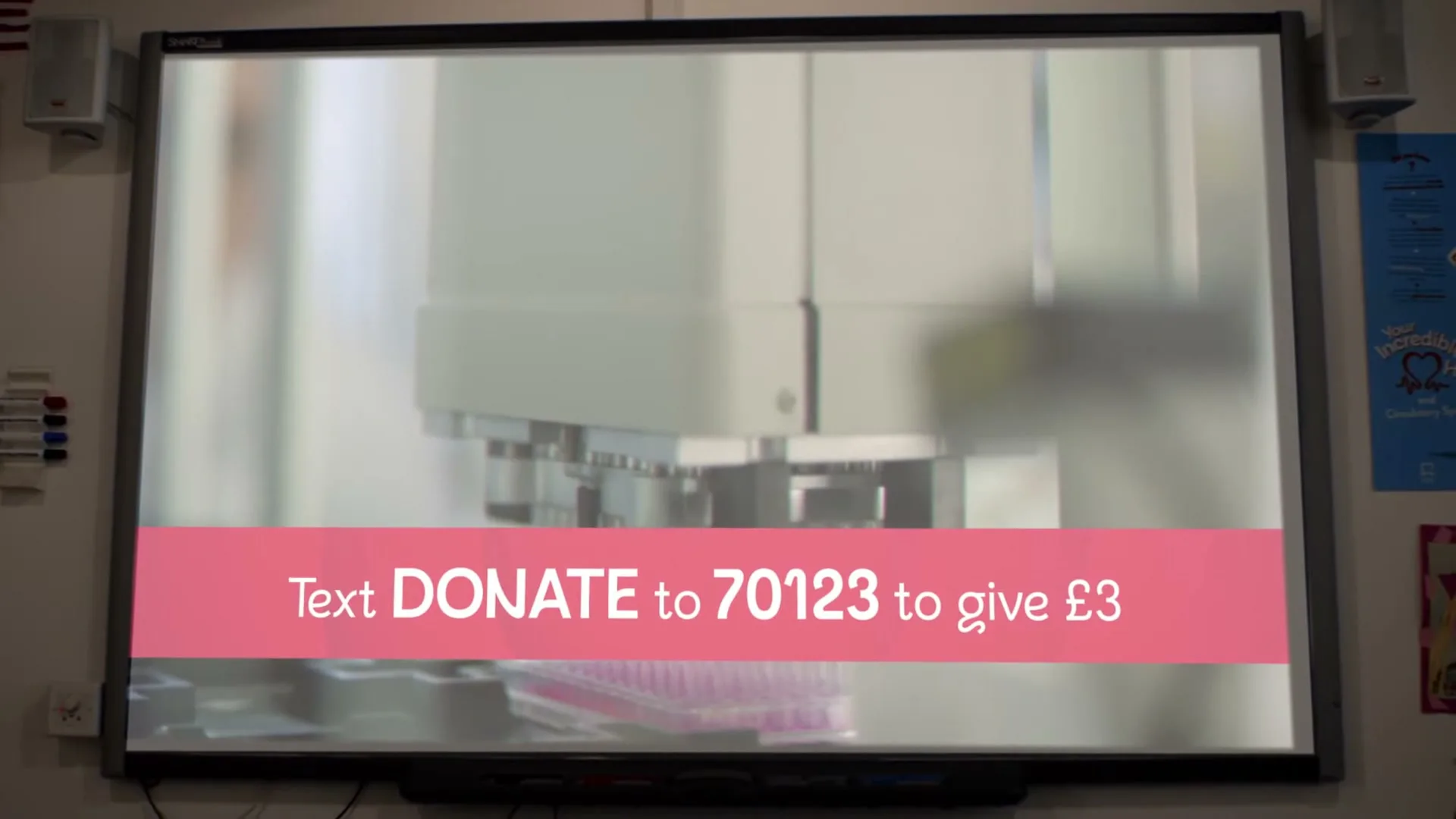 A wide shot of a classroom whiteboard displaying a video of scientific equipment and a pink banner with donation instructions.