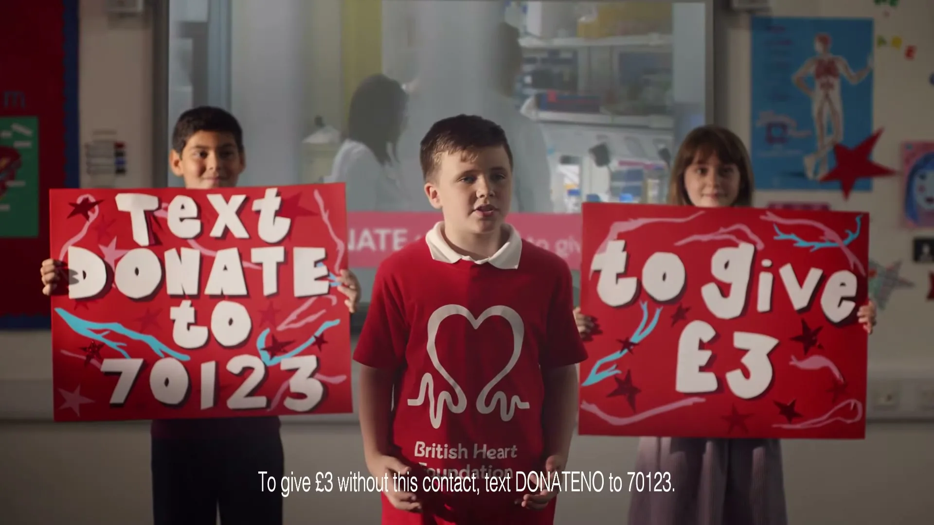 Three children in a classroom hold up signs asking for donations to the British Heart Foundation.