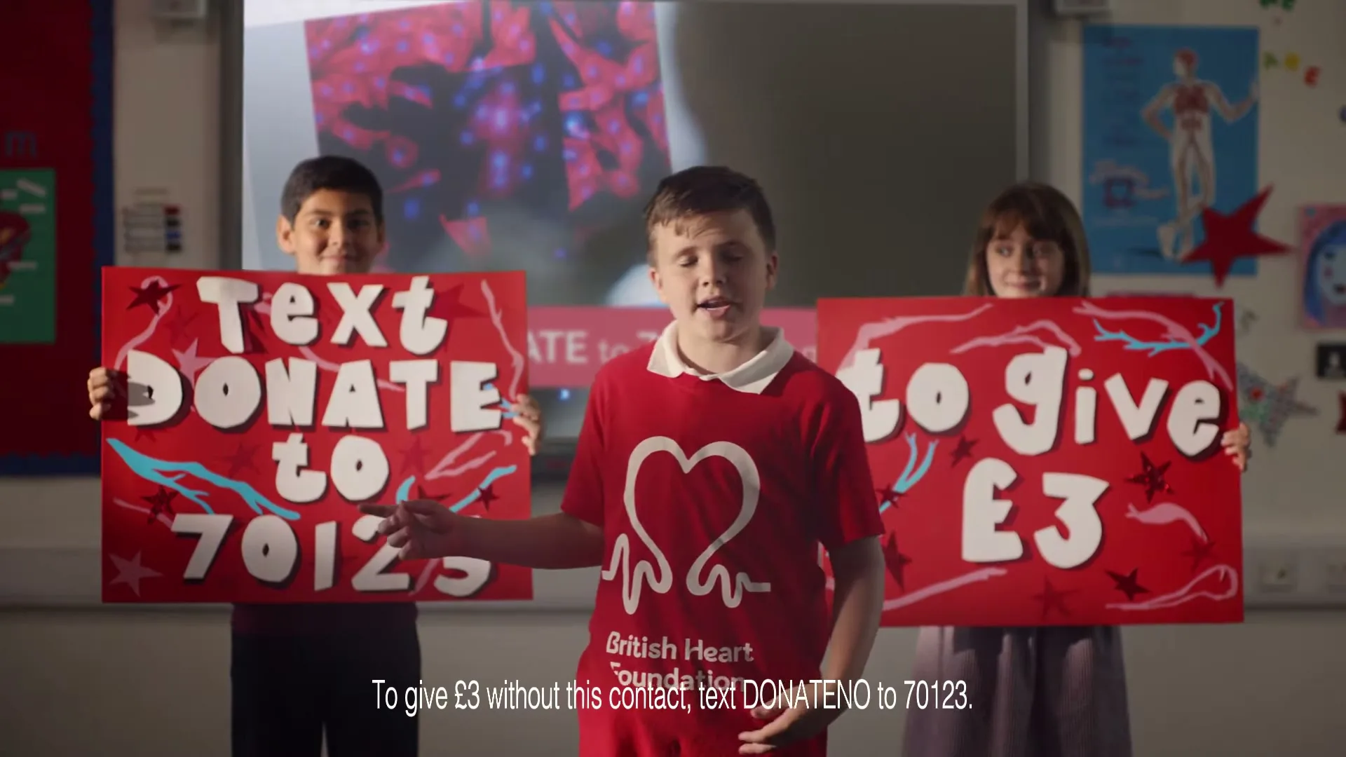 Three children in a classroom hold up red signs encouraging viewers to text to donate money to the British Heart Foundation.