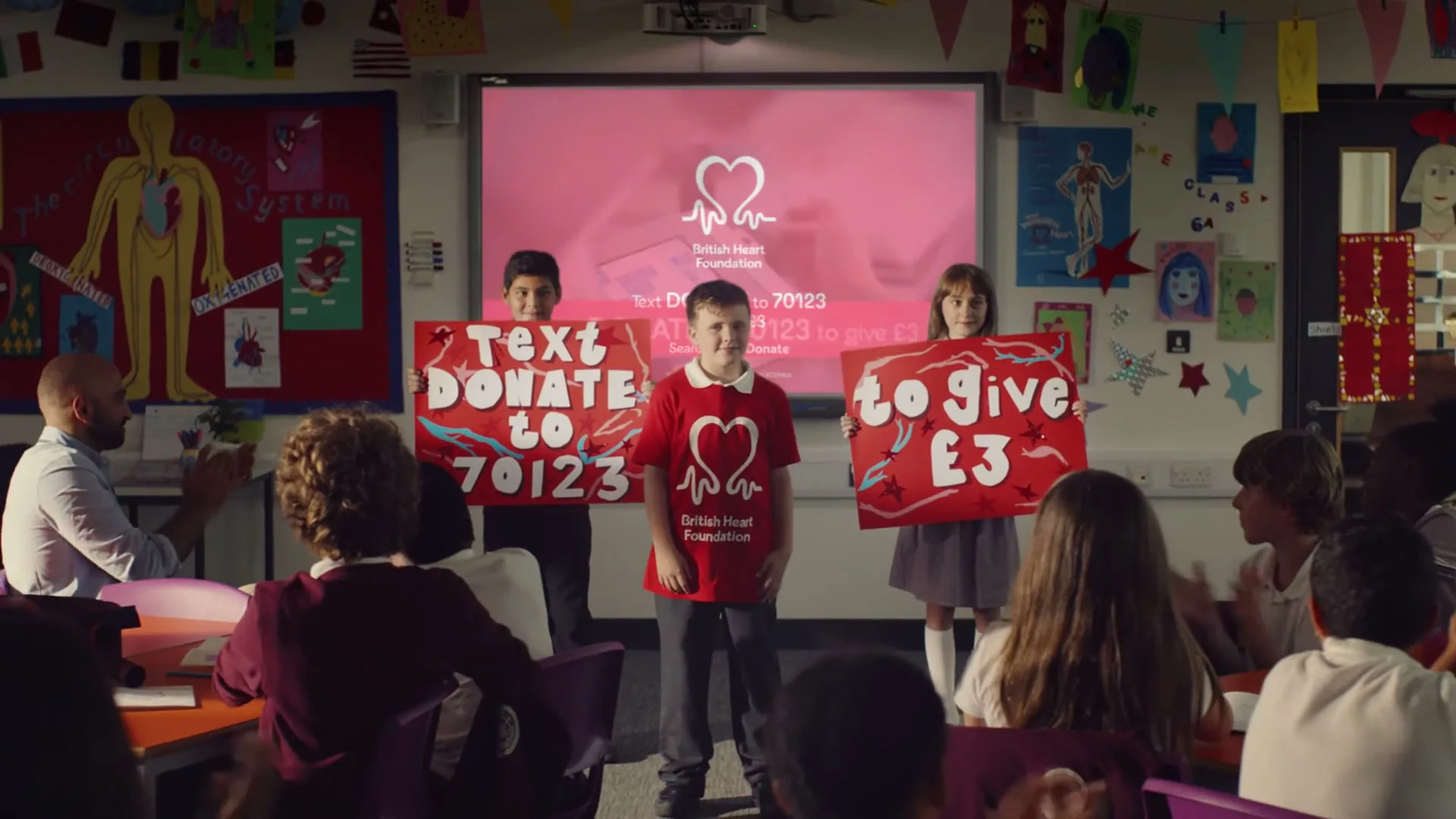 A young boy in a red t-shirt stands in front of a classroom, holding a sign, flanked by two other children holding signs, as classmates applaud.