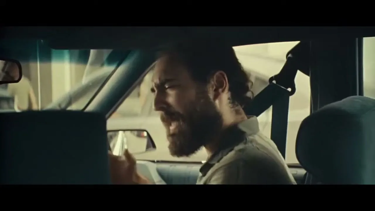 A bearded man in his 30s, wearing a polo shirt, argues with someone off-screen while driving a car during the day.