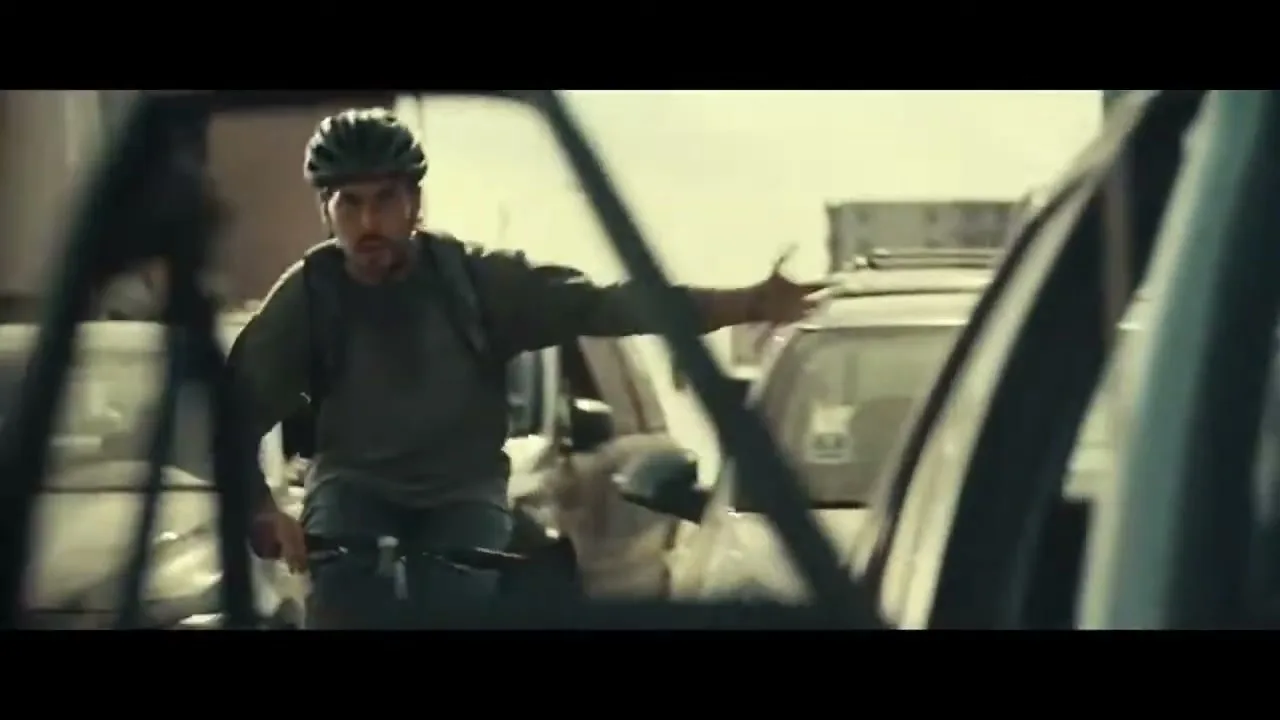A male cyclist in a helmet and backpack rides through heavy traffic, looking frustrated and gesturing with his hand, seen through a car window.