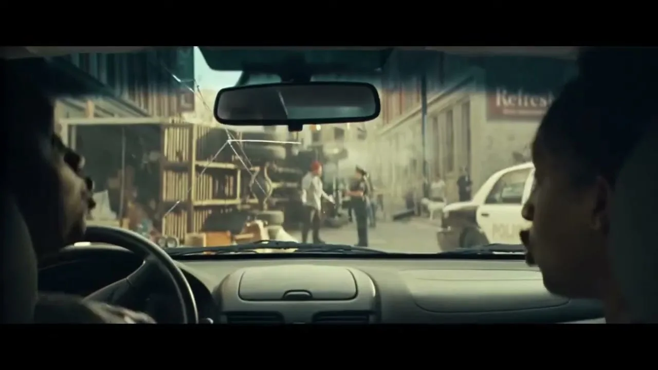 Two people are seen from behind inside a car, looking out the windshield at a street scene with people and a police car.