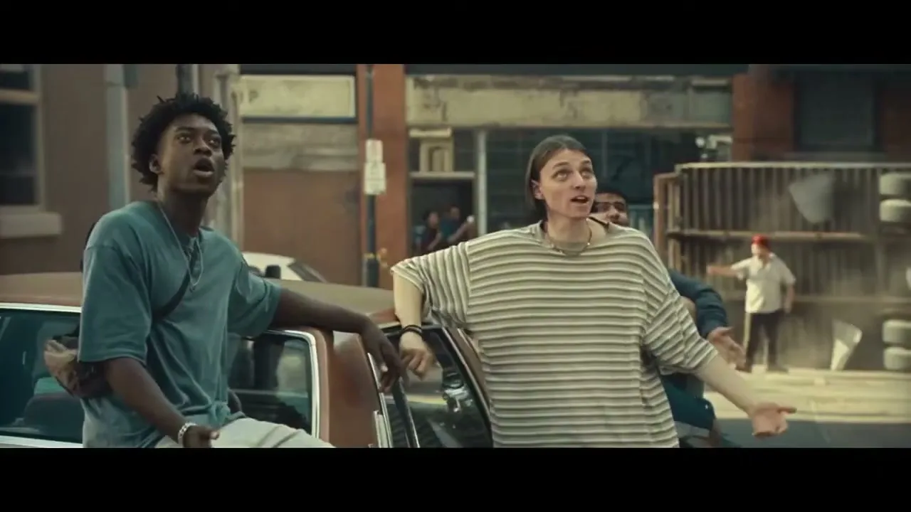 Two young people, a Black male and a white female, lean on a brown car, looking up in surprise on a city street.