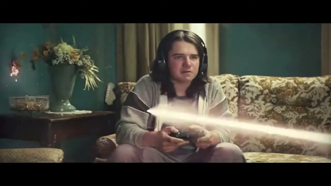 A young person with long hair and headphones plays a video game on a patterned couch in a dimly lit living room, reacting to an on-screen event.