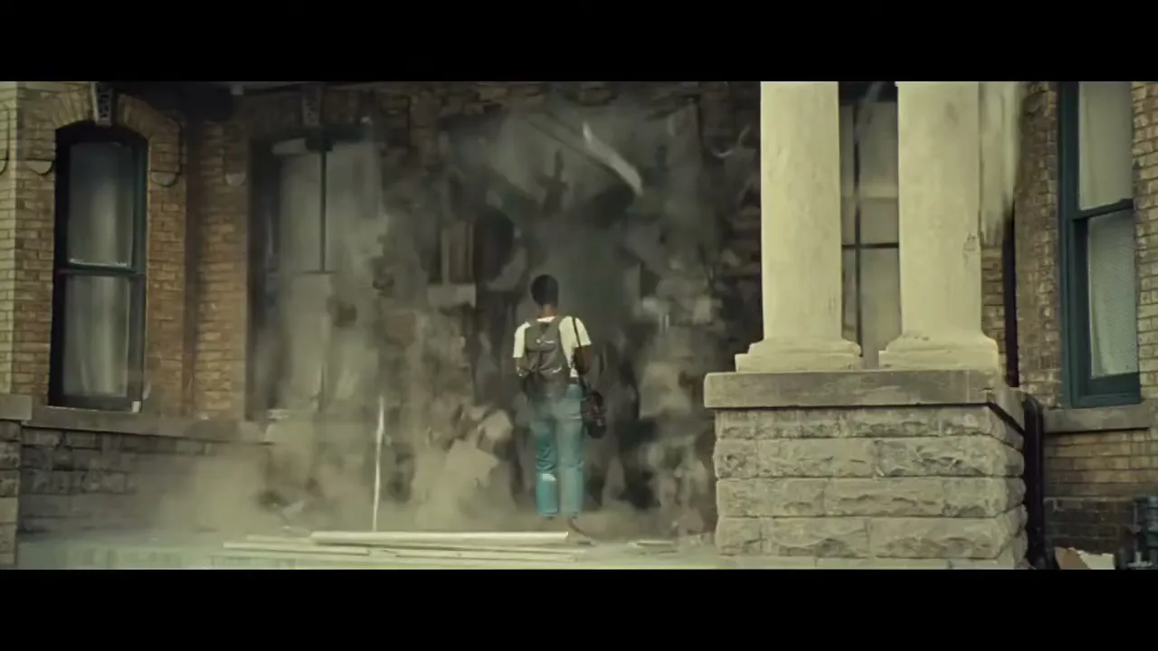 A young woman with a backpack stands in front of a brick building as the wall behind her explodes, creating a cloud of dust and debris.