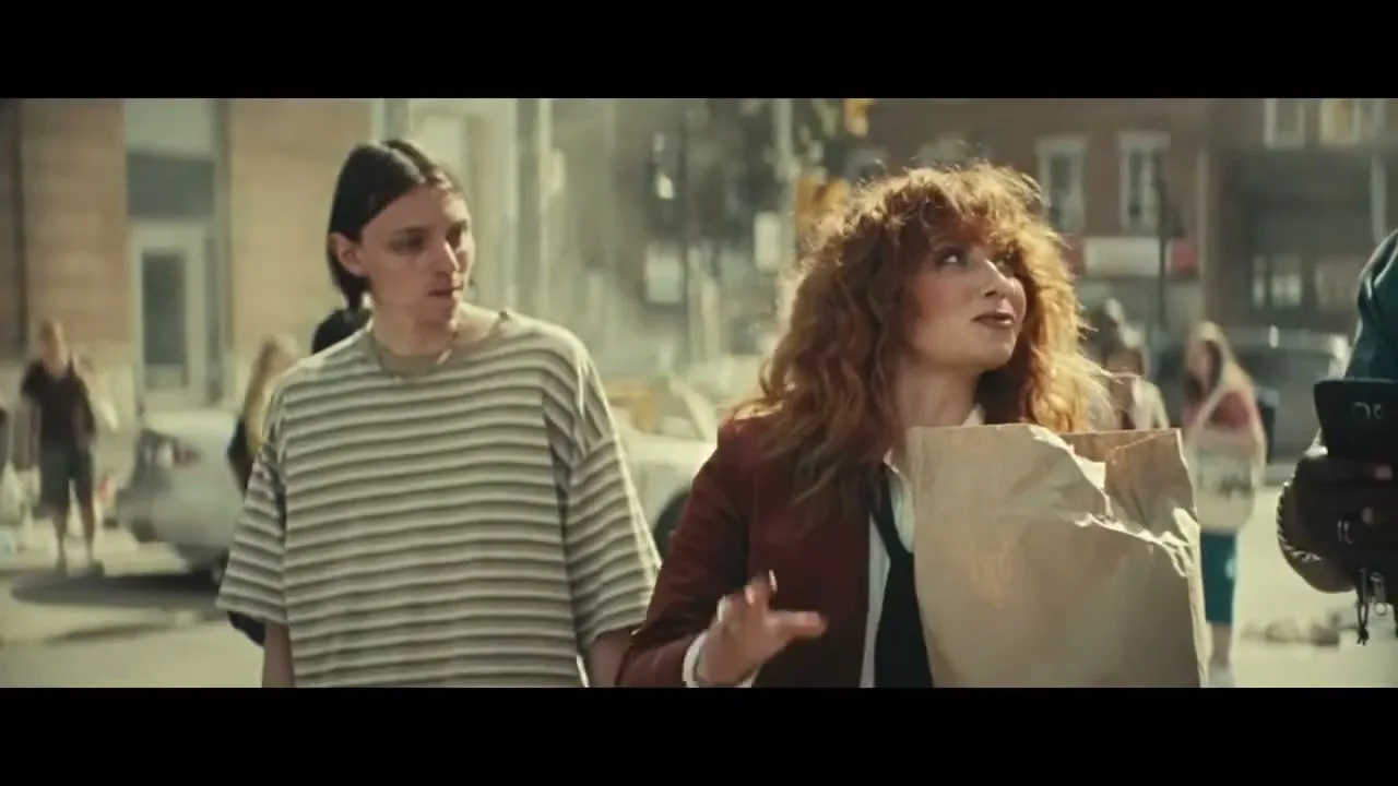 A woman with red curly hair holds a paper bag and looks up, while a man in a striped shirt stands beside her on a city street.