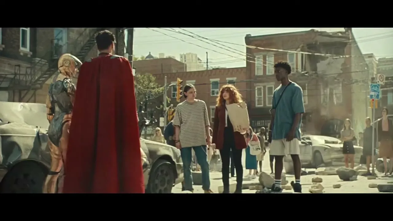 A diverse group of young people stands on a debris-filled street, looking at a man in a red cape and a golden robot, in a post-apocalyptic city.