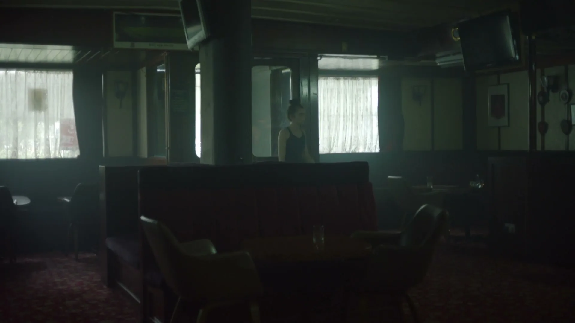 A young woman with dark hair in a bun stands in a dimly lit, empty bar or lounge area, looking directly at the camera.