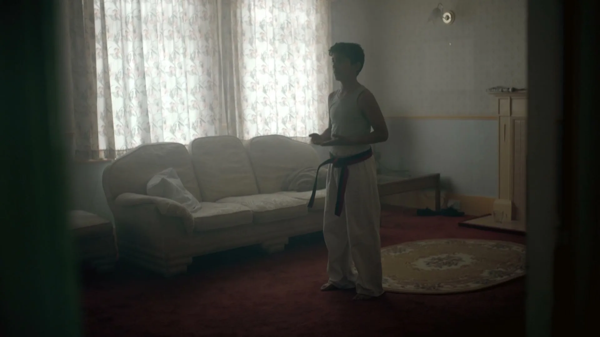 A young boy in a white karate gi with a black belt stands in a dimly lit living room, practicing his moves.