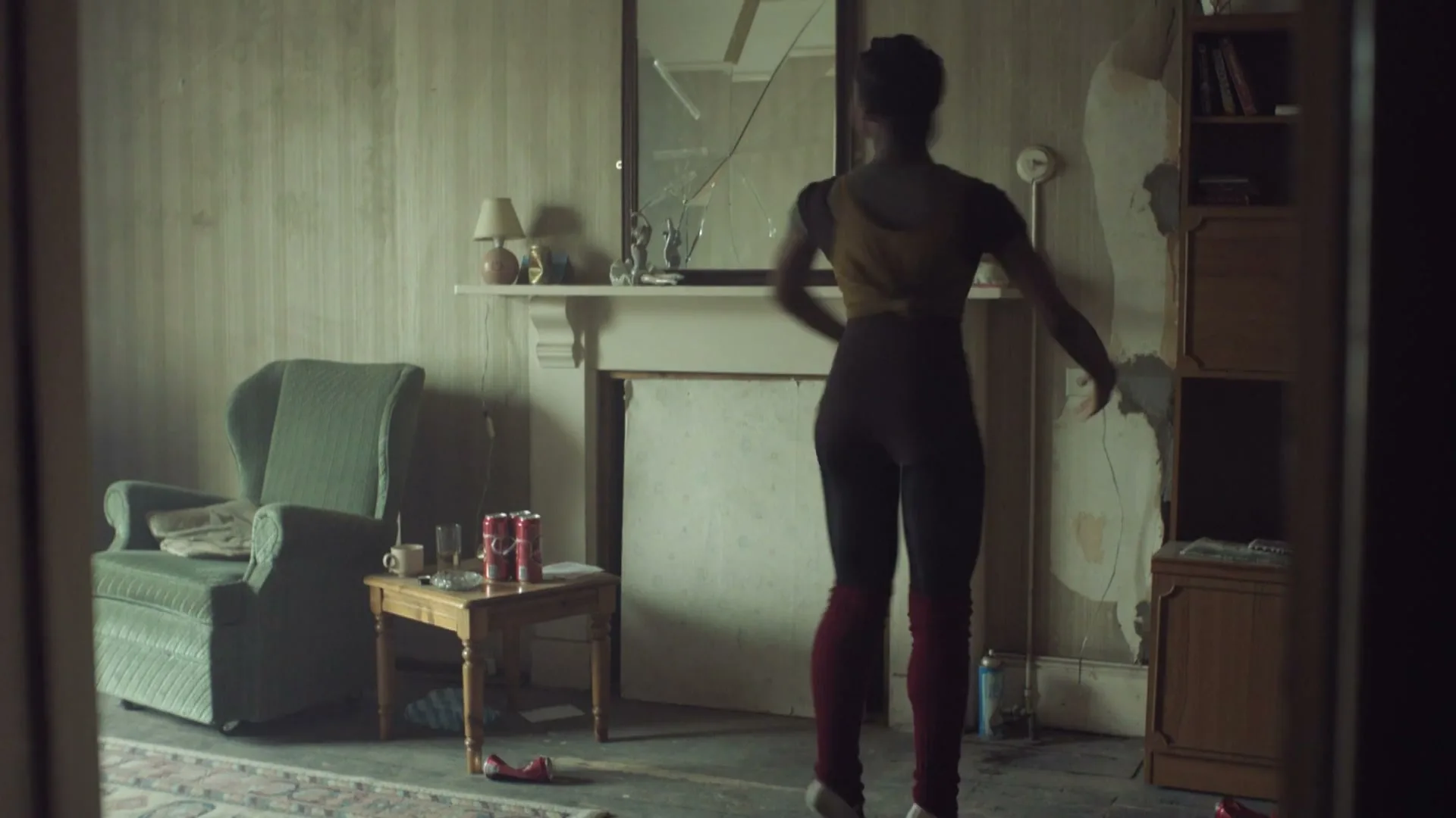 A young Black woman in workout clothes dances in a dilapidated living room with a broken mirror over the fireplace.