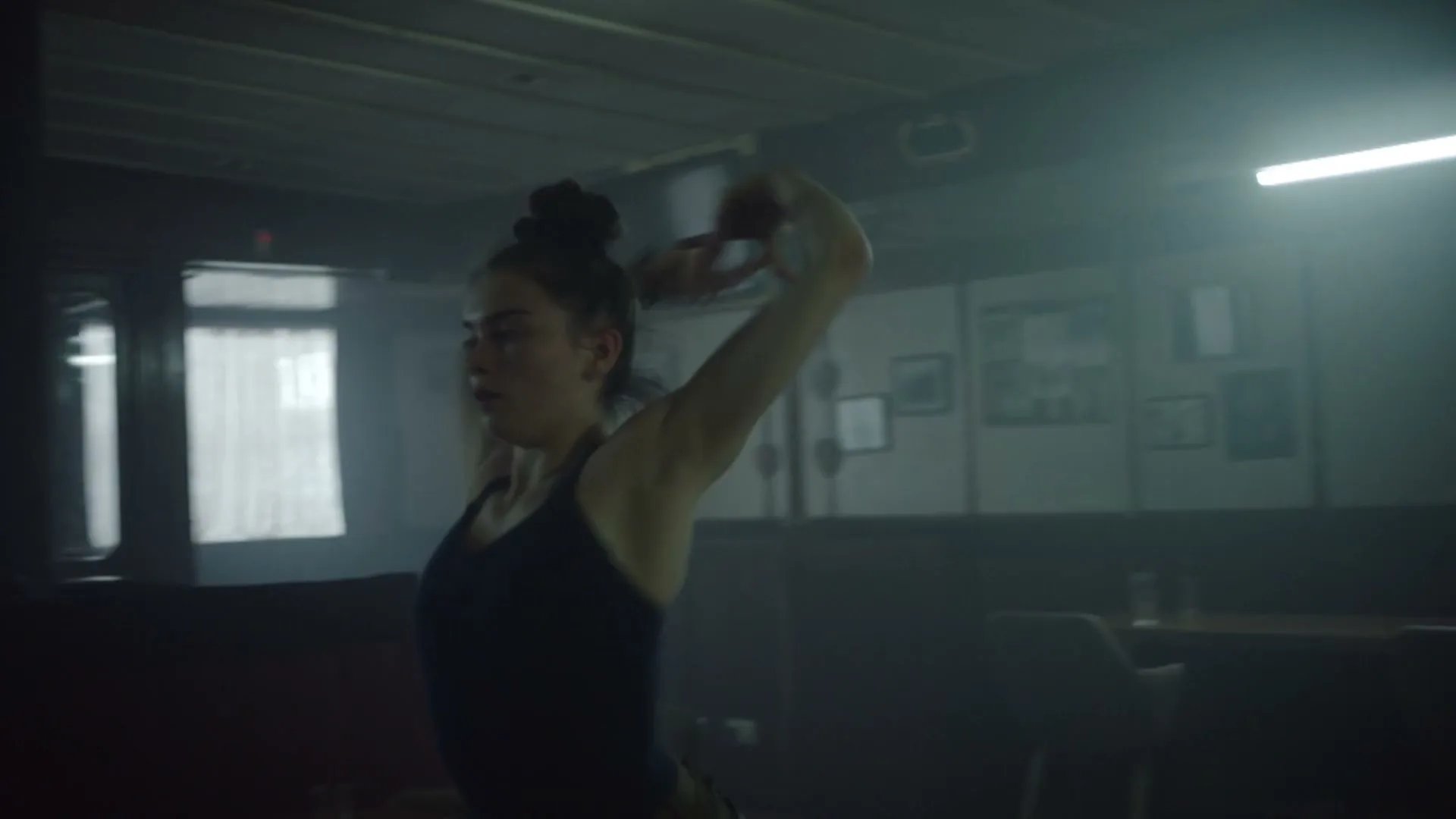 A young woman with her hair in a bun, wearing a dark tank top, dances expressively in a dimly lit, dusty room with windows and tables.