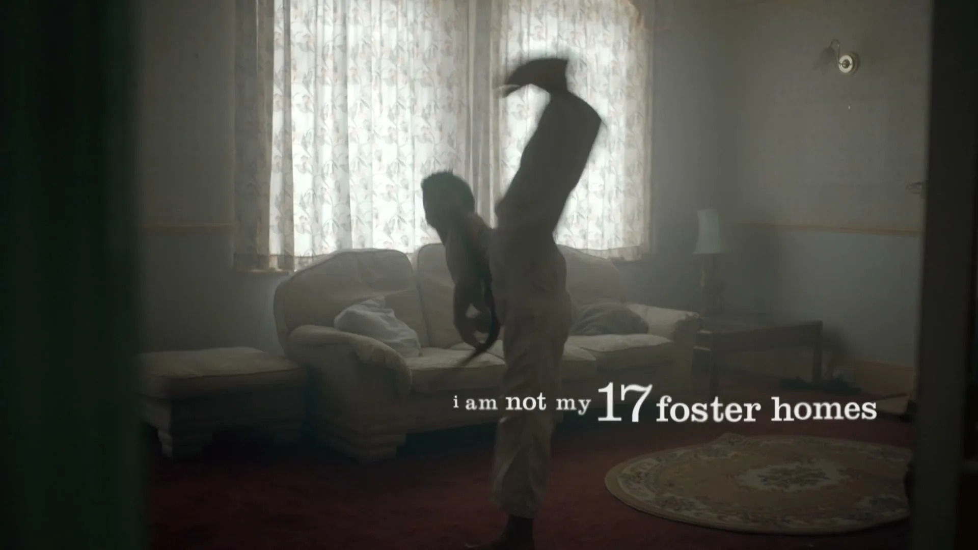 A young male in a white martial arts uniform practices a kick in a dimly lit living room with patterned curtains and a sofa.