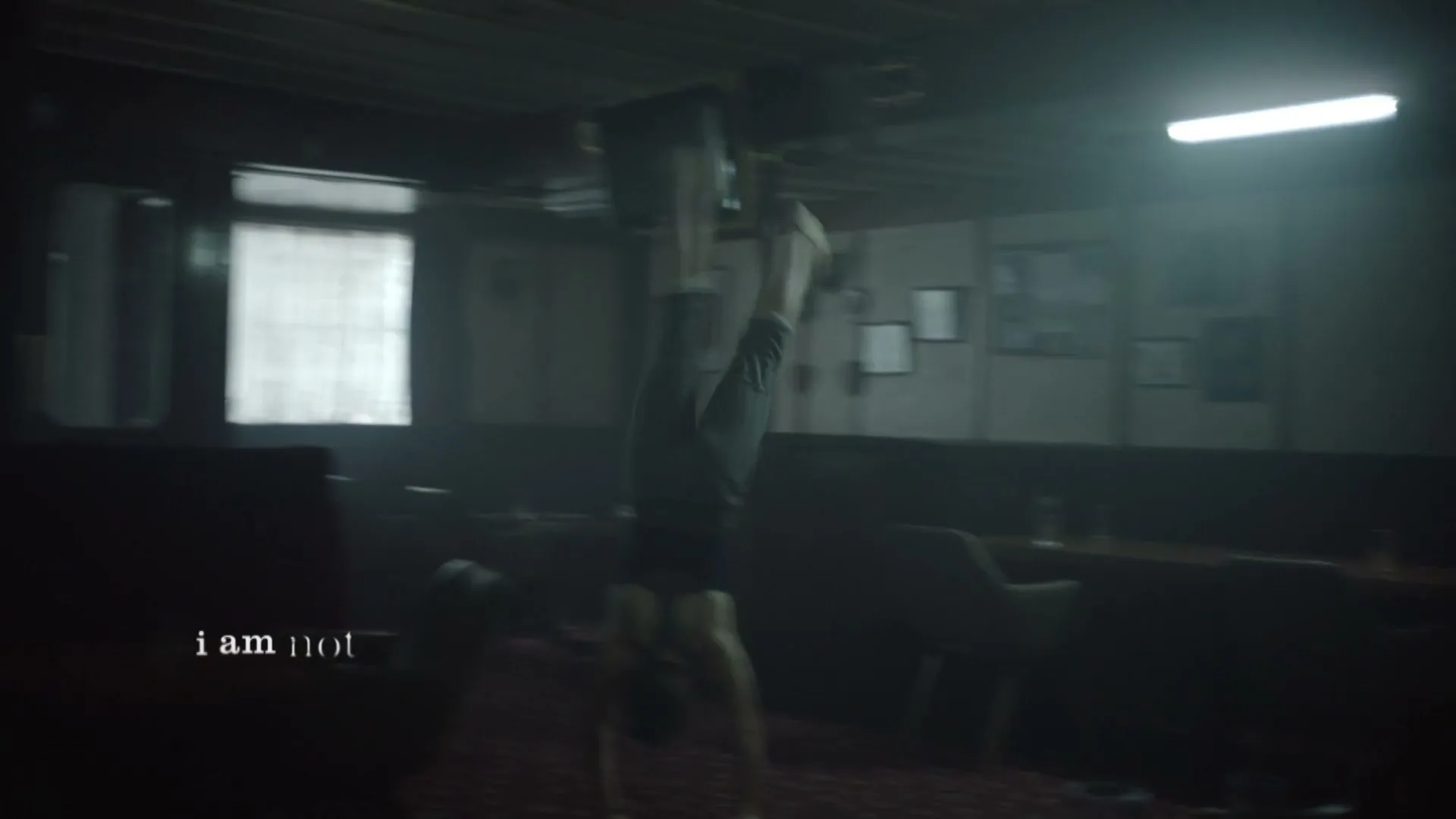A young woman in a dark room with windows and tables does a handstand, then falls, as text appears on screen.