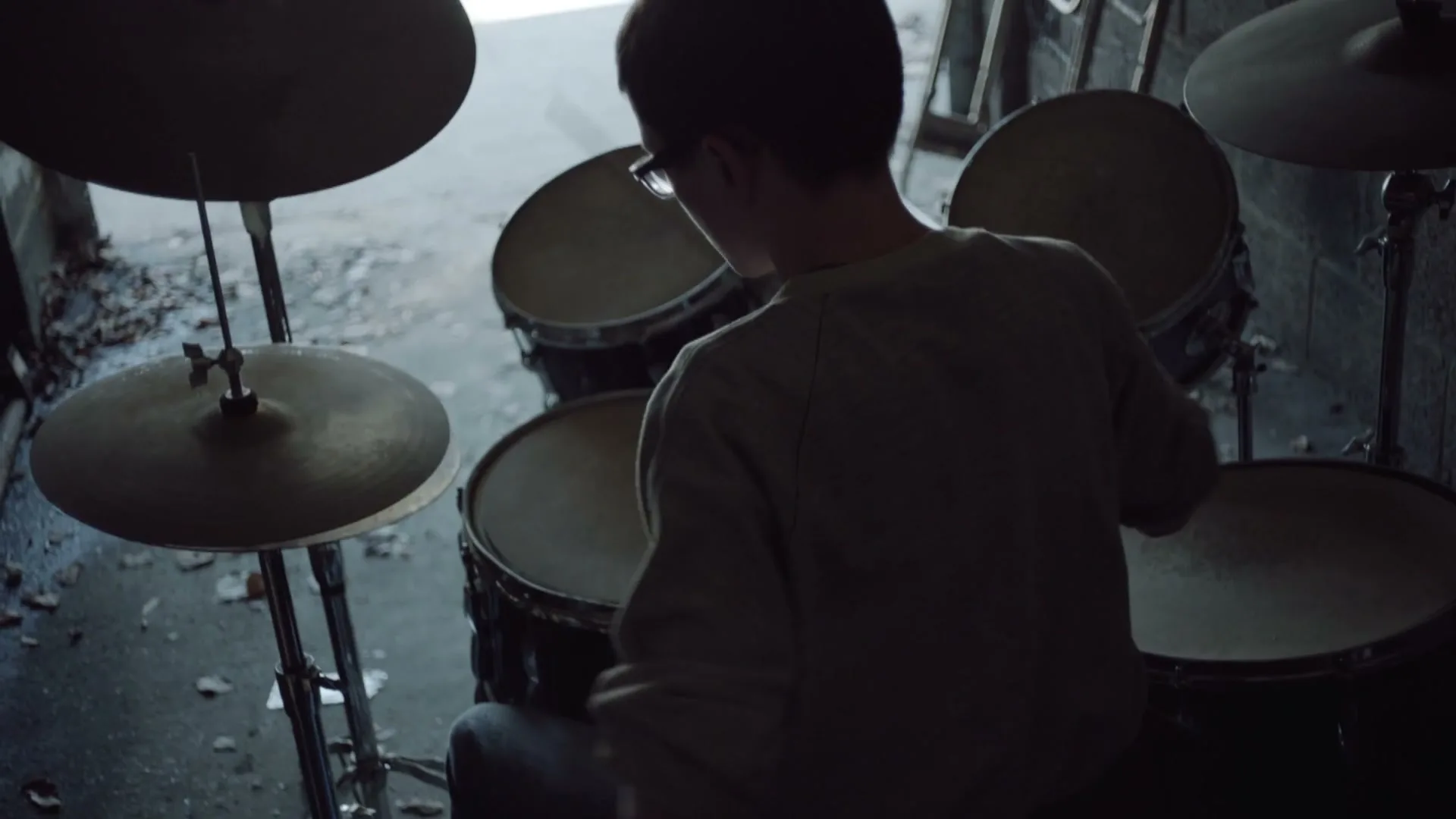 A young male in glasses and a sweatshirt plays drums in a dark, gritty garage, with the word "worthless" appearing on screen.