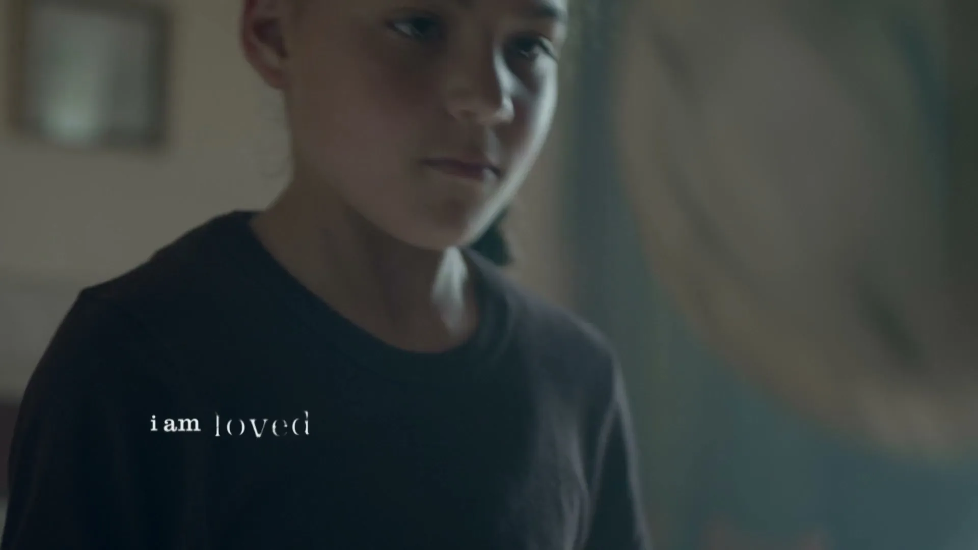 A young girl in a dark t-shirt looks down, with the words "i am loved" appearing on screen, in a dimly lit room.