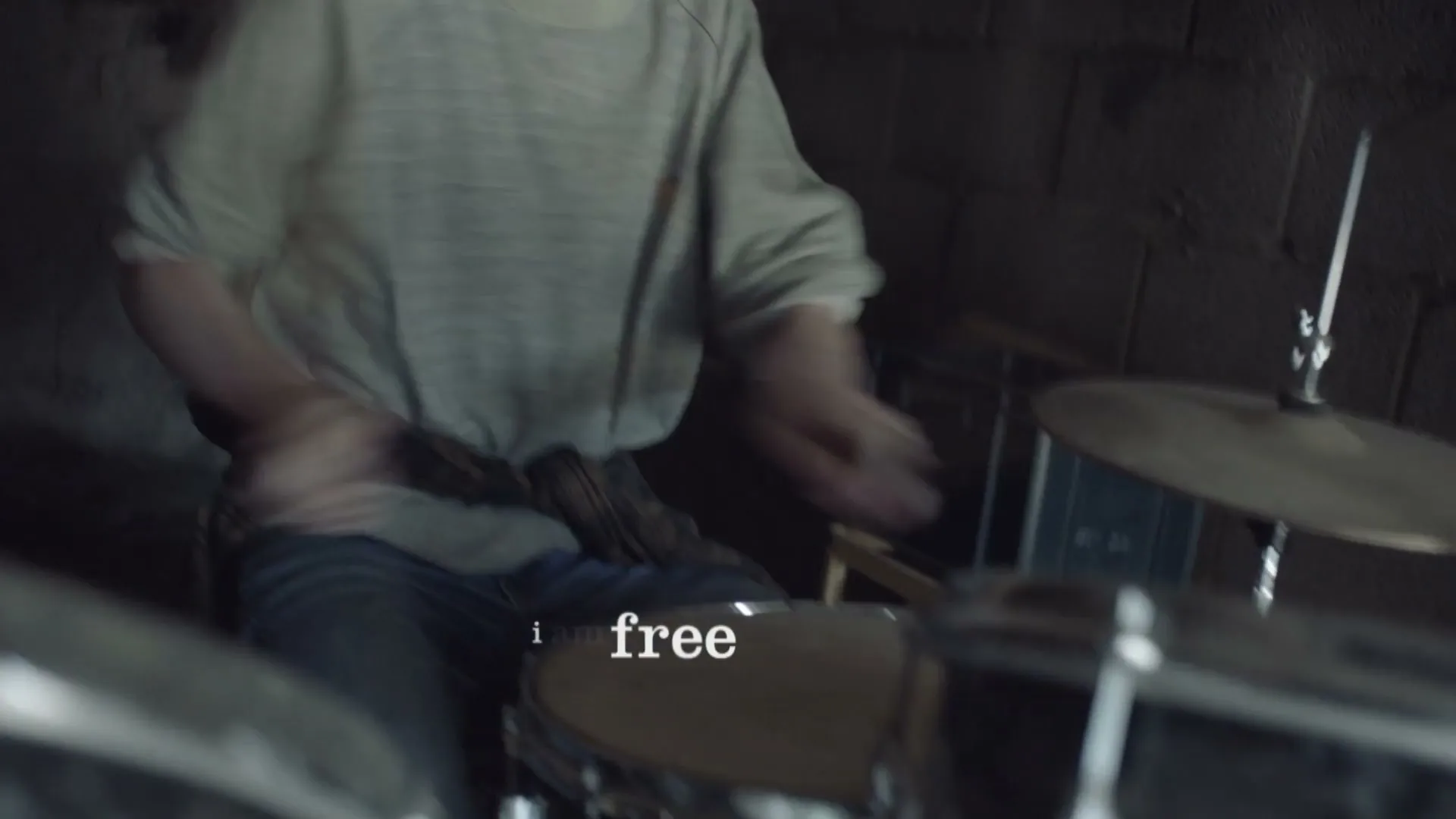 A young person with glasses and a grey shirt plays drums in a dimly lit room, with the text "i am free" on screen.