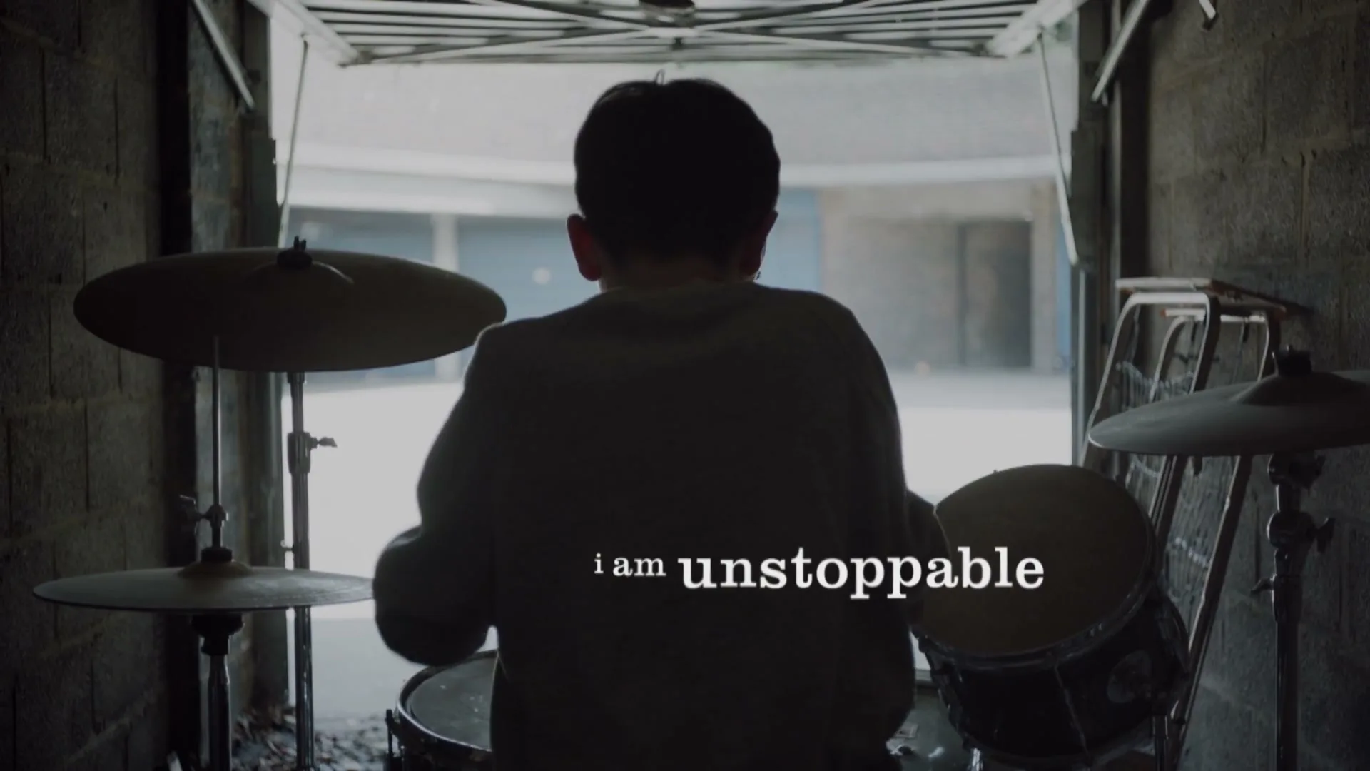A young boy with dark hair, wearing a light-colored sweatshirt, sits at a drum kit in a dimly lit garage, facing away from the camera.