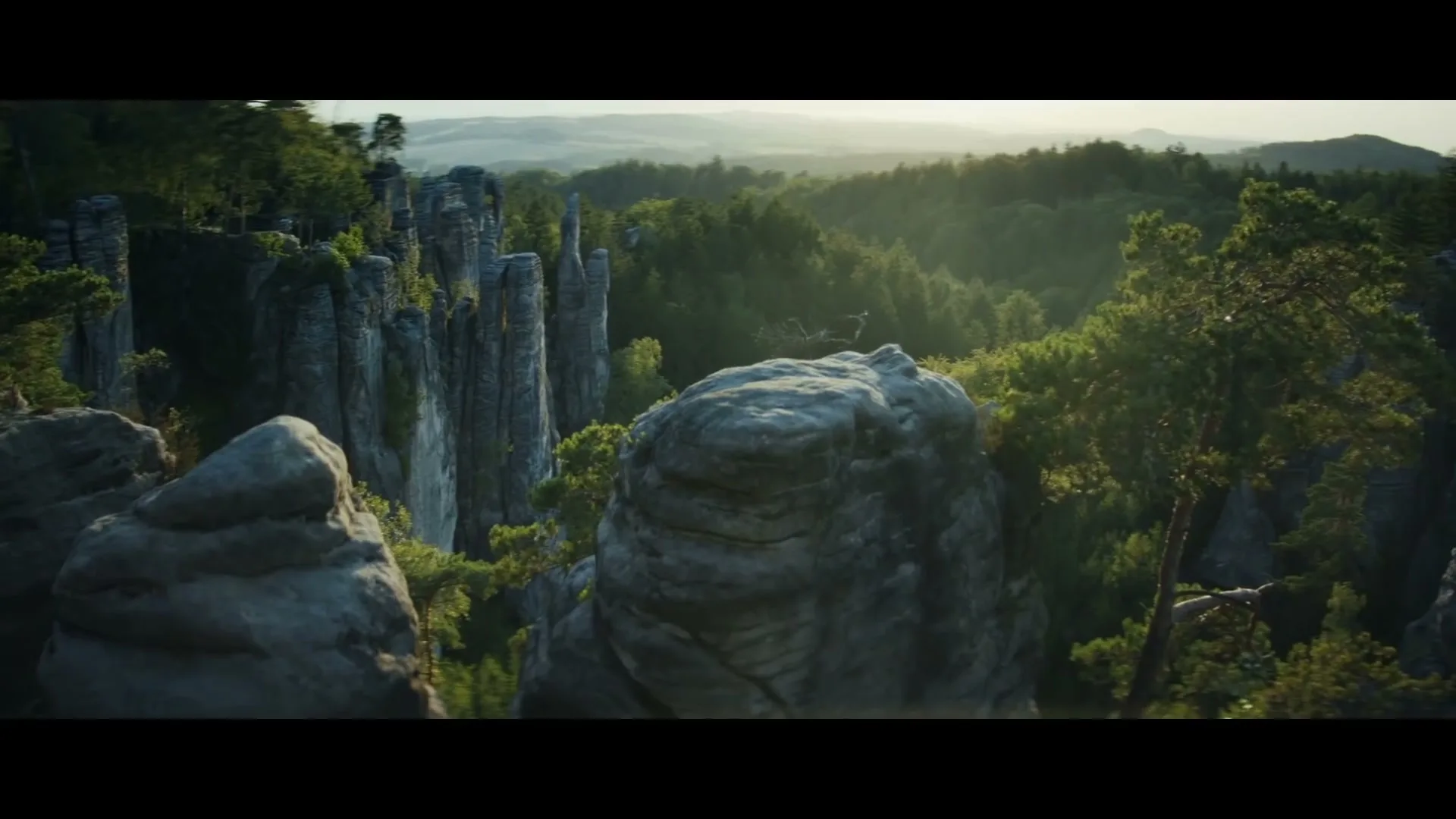 An aerial shot captures a vast, lush green forest with towering rock formations under a bright sky, with sunlight filtering through the trees.
