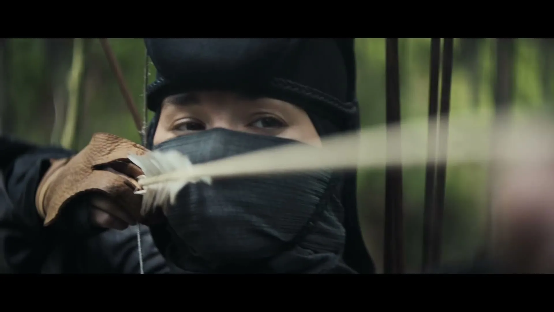 A young female archer, dressed in black and wearing a face covering, aims her bow and arrow in a forest.