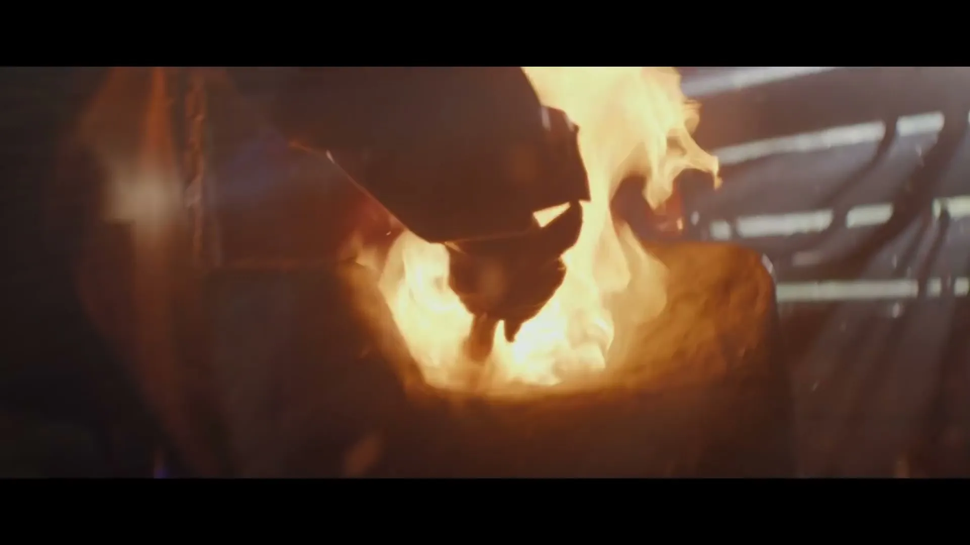 A blacksmith's hand holds a metal object, heating it in a fiery forge, with light rays shining through wooden slats in a dark workshop.