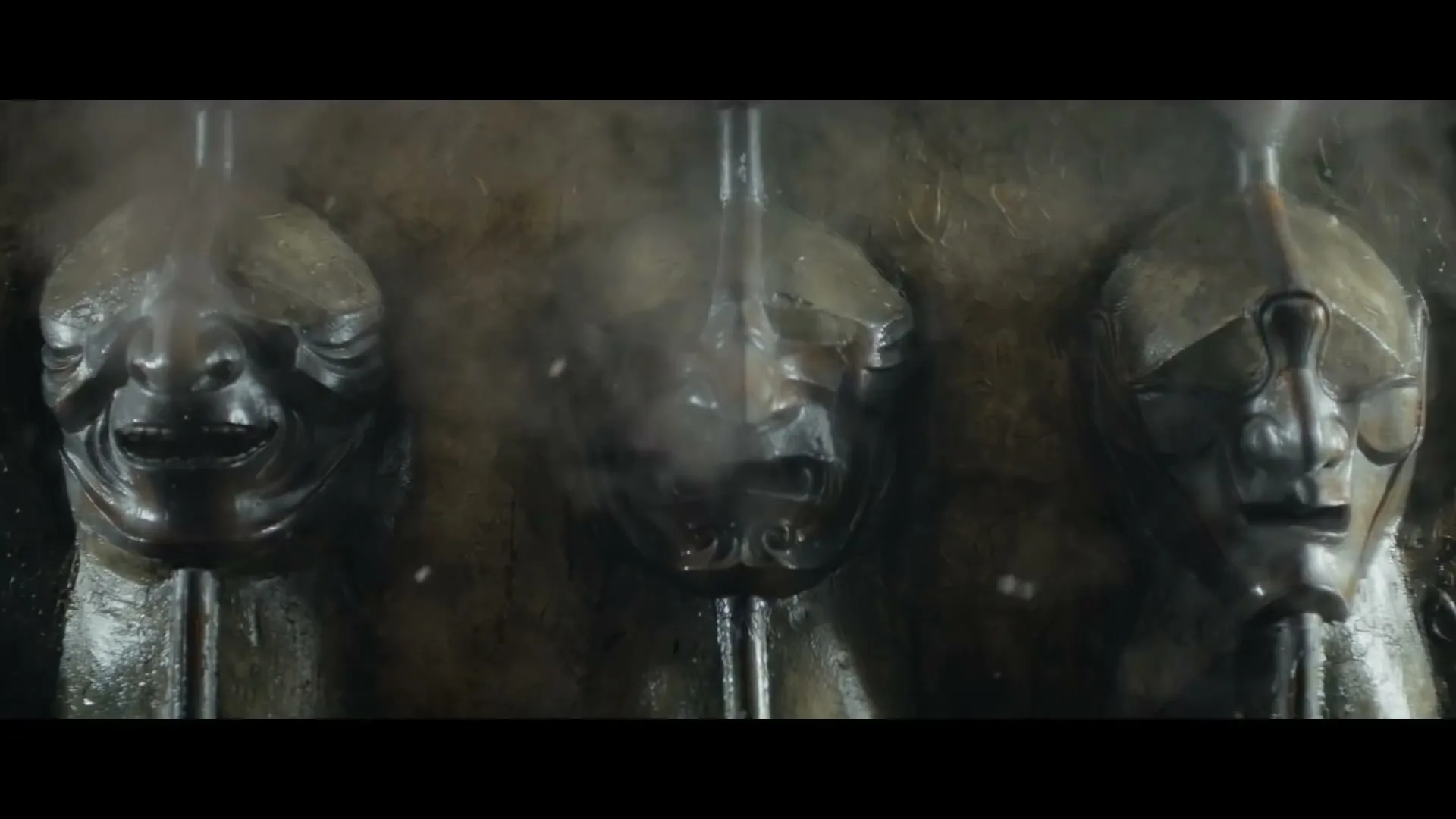 Three ancient, weathered metal masks with distinct expressions are seen in a row, with steam rising around them in a dark, mysterious setting.