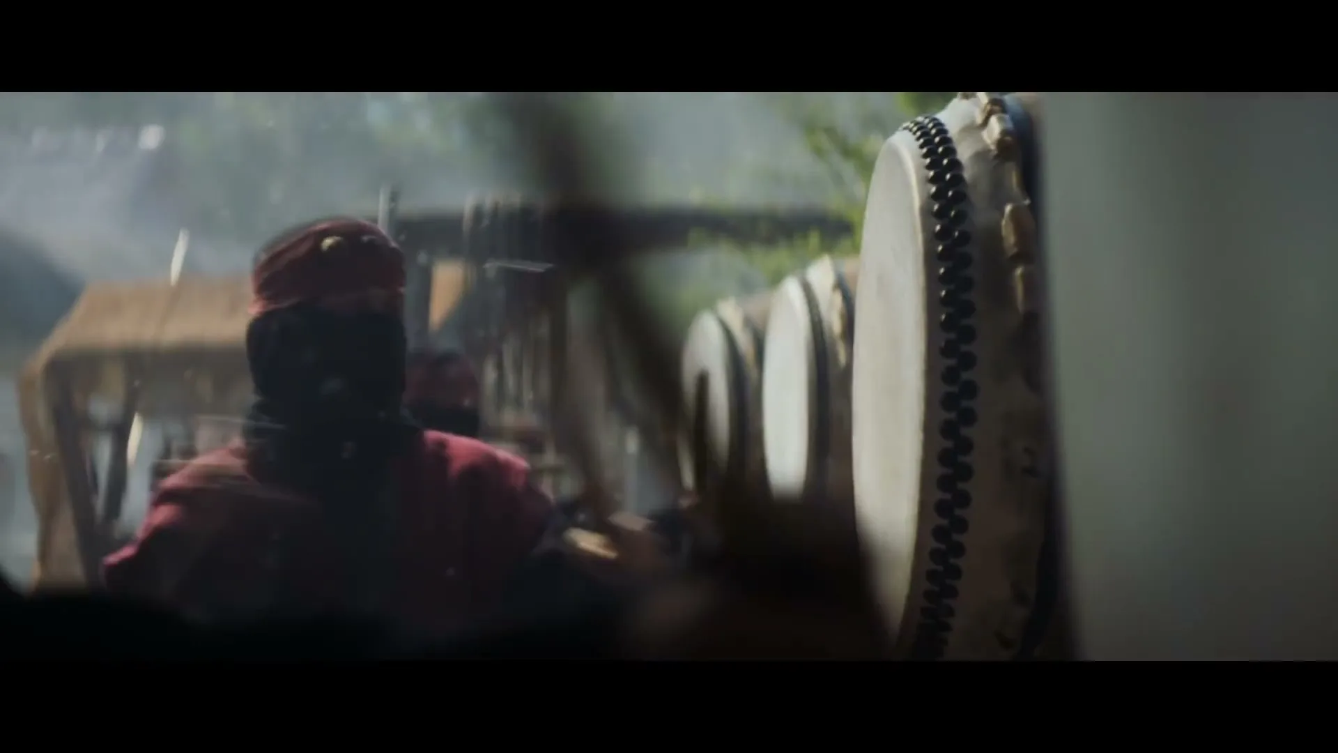 A person in traditional Japanese attire, with a red headband and black face covering, vigorously plays a large taiko drum outdoors.