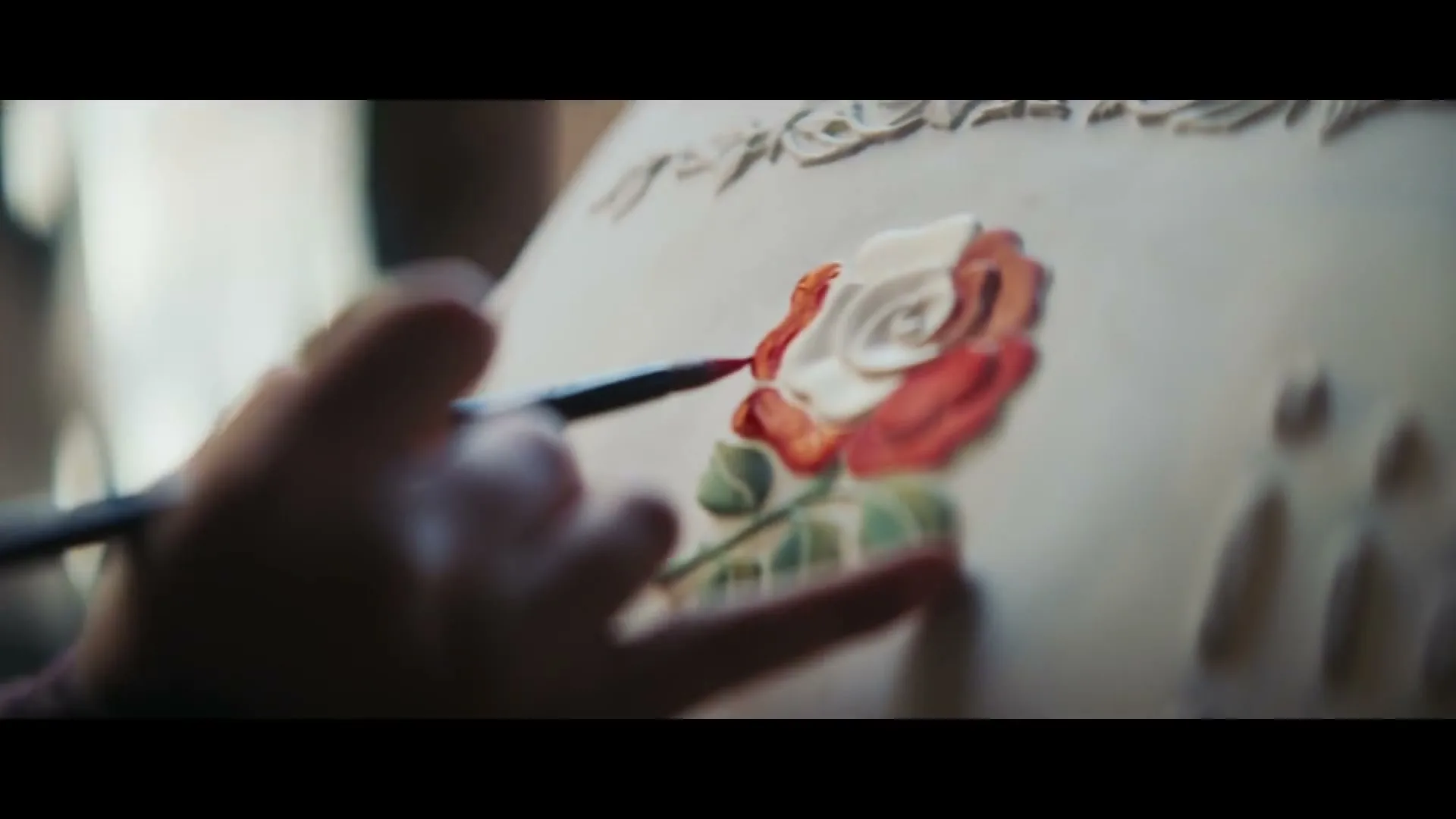 A person's hand delicately paints a red and white rose onto a white surface, possibly a cake or ceramic, in a close-up shot.