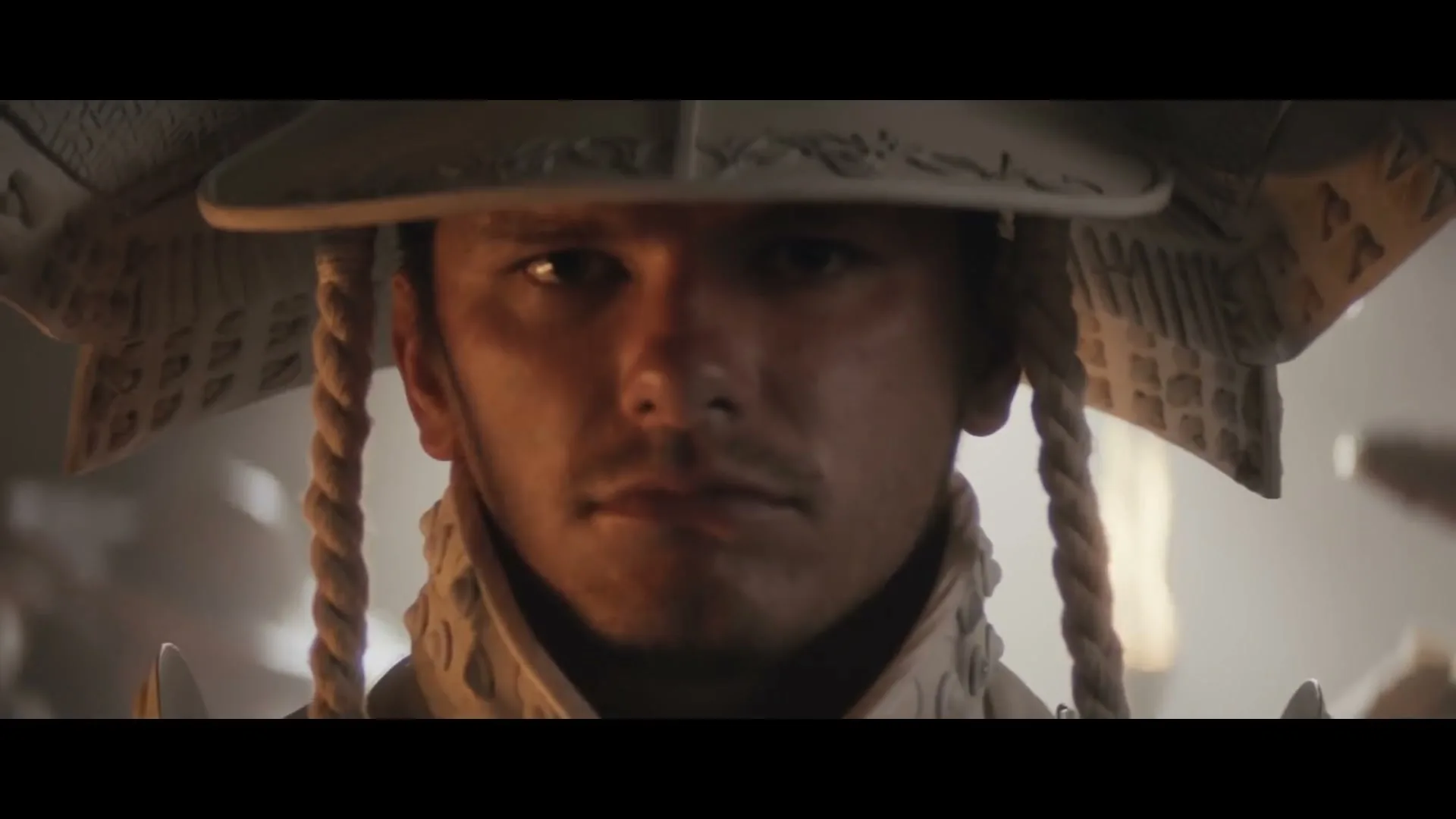A young man in white samurai armor looks directly at the camera as two hands adjust his helmet.