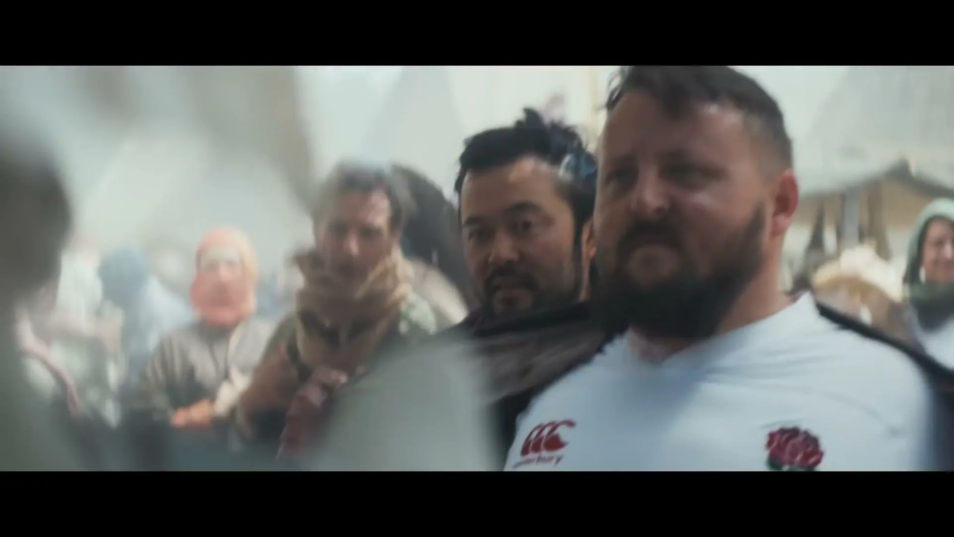 A male rugby player in a white jersey stands among a crowd of people, looking determined in a dusty, chaotic environment.