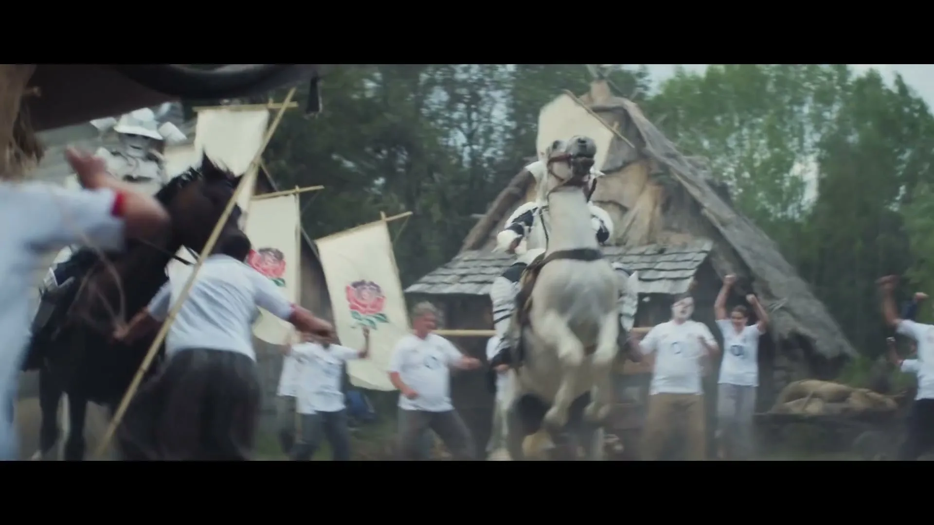 A knight in shining armor on a white horse rears up in a medieval village, surrounded by cheering people in white t-shirts.
